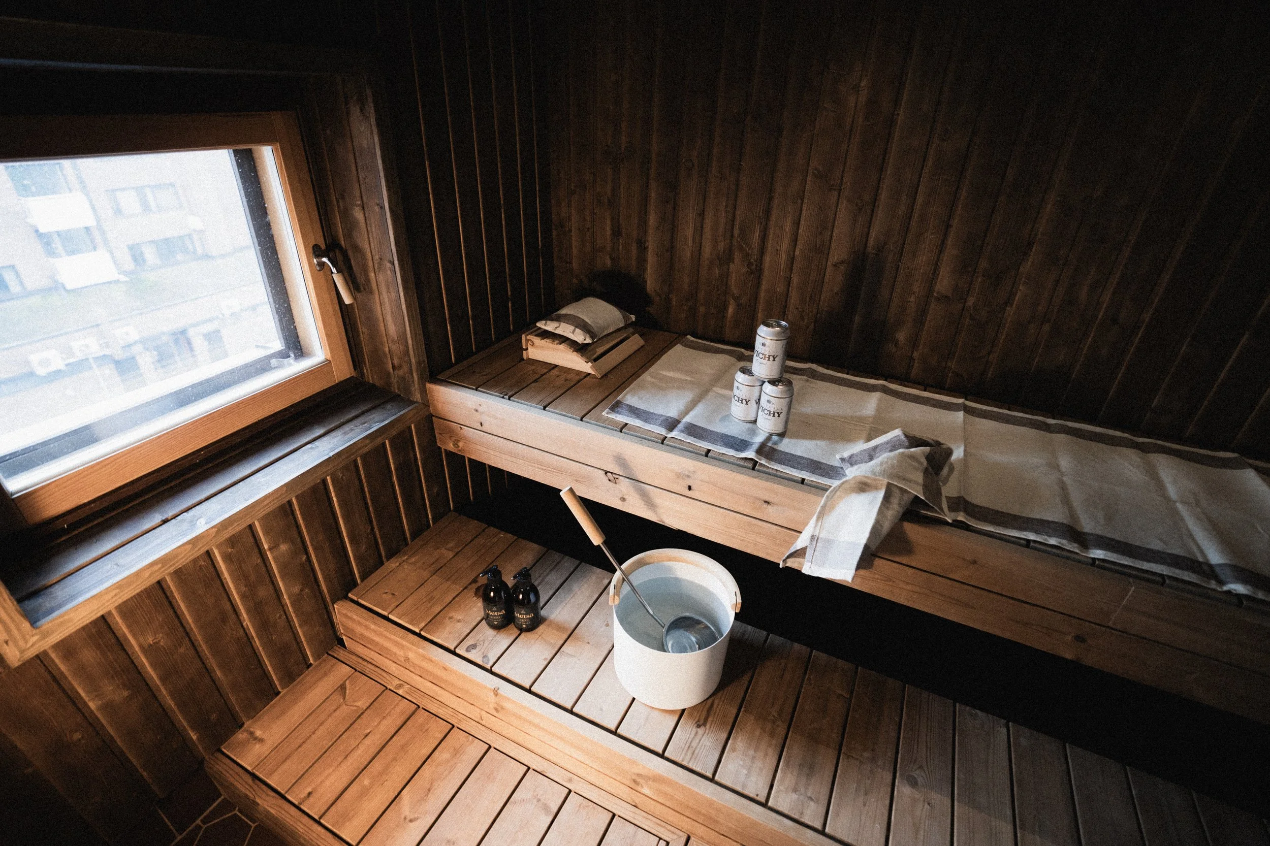Sauna in Rovaniemi. Cozy wooden sauna room with a window, a bench with a white towel, three cans of beer, a pillow, a stack of books, a bucket with a ladle, and two bottles of soap or shampoo.