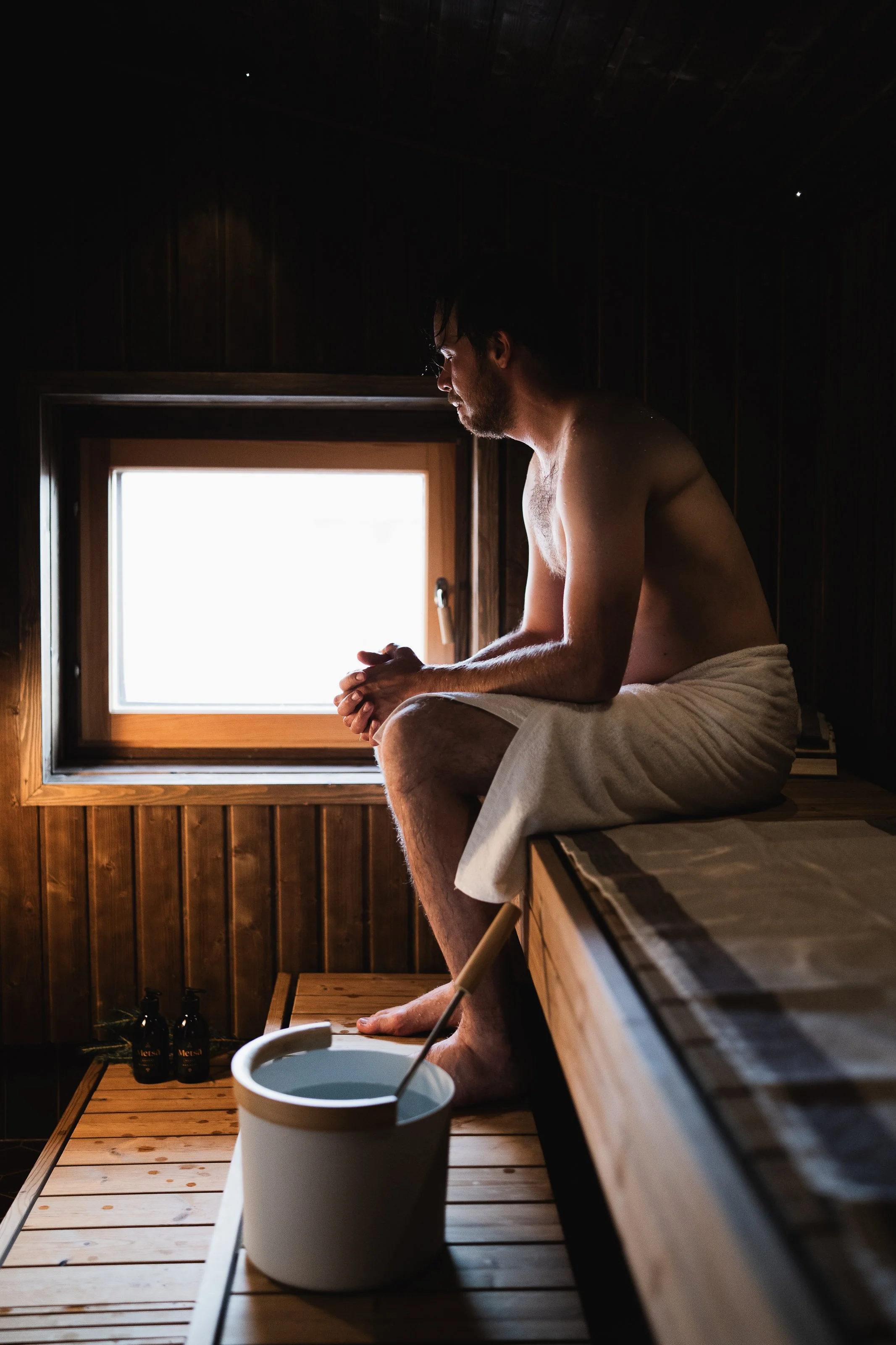 A man sitting on a wooden bench in a sauna, wrapped in a towel, looking out a small window, with sauna bottles and a bucket nearby.
