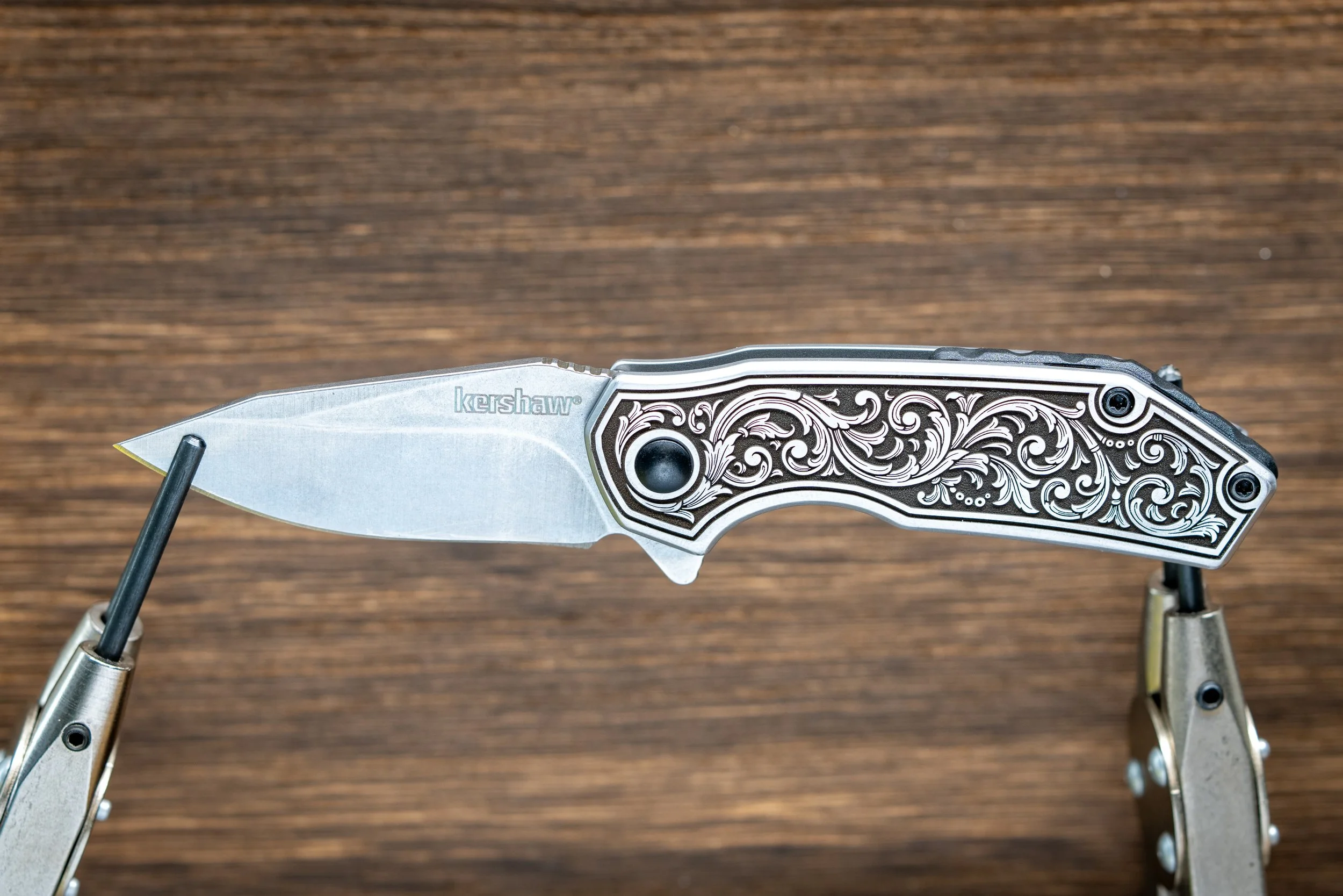 A folding pocket knife with a silver blade and a decorative handle with intricate floral engravings, held open by black and silver mechanical arms on a wooden surface.