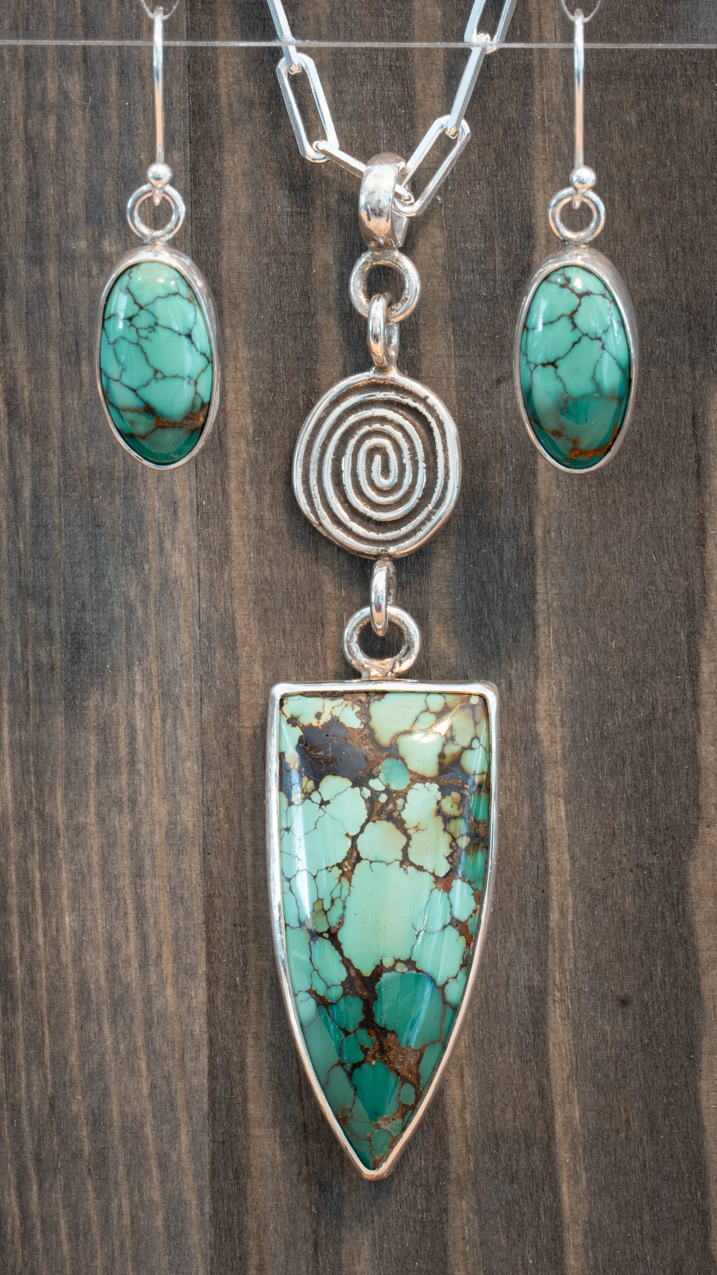 Three jewelry pieces featuring turquoise stones on a wooden background. The necklace has an oval turquoise pendant, the earrings have matching oval turquoise stones, and the pendant has a large irregular turquoise stone with brown veining.