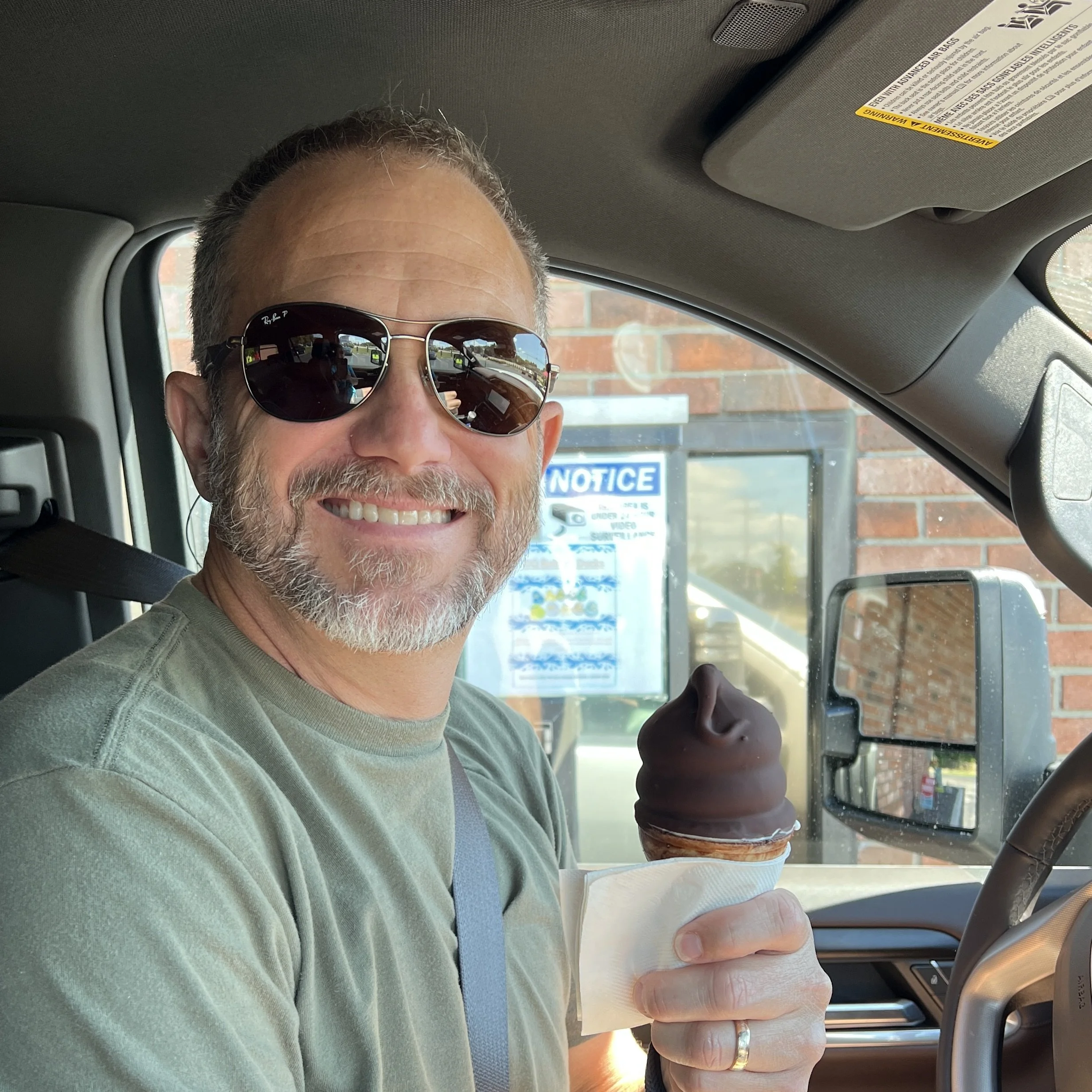 A man wearing sunglasses and a green shirt sitting inside a vehicle, holding an ice cream cone with chocolate ice cream. He is smiling and looking at the camera. The background shows a brick wall and a notice sign outside the vehicle.