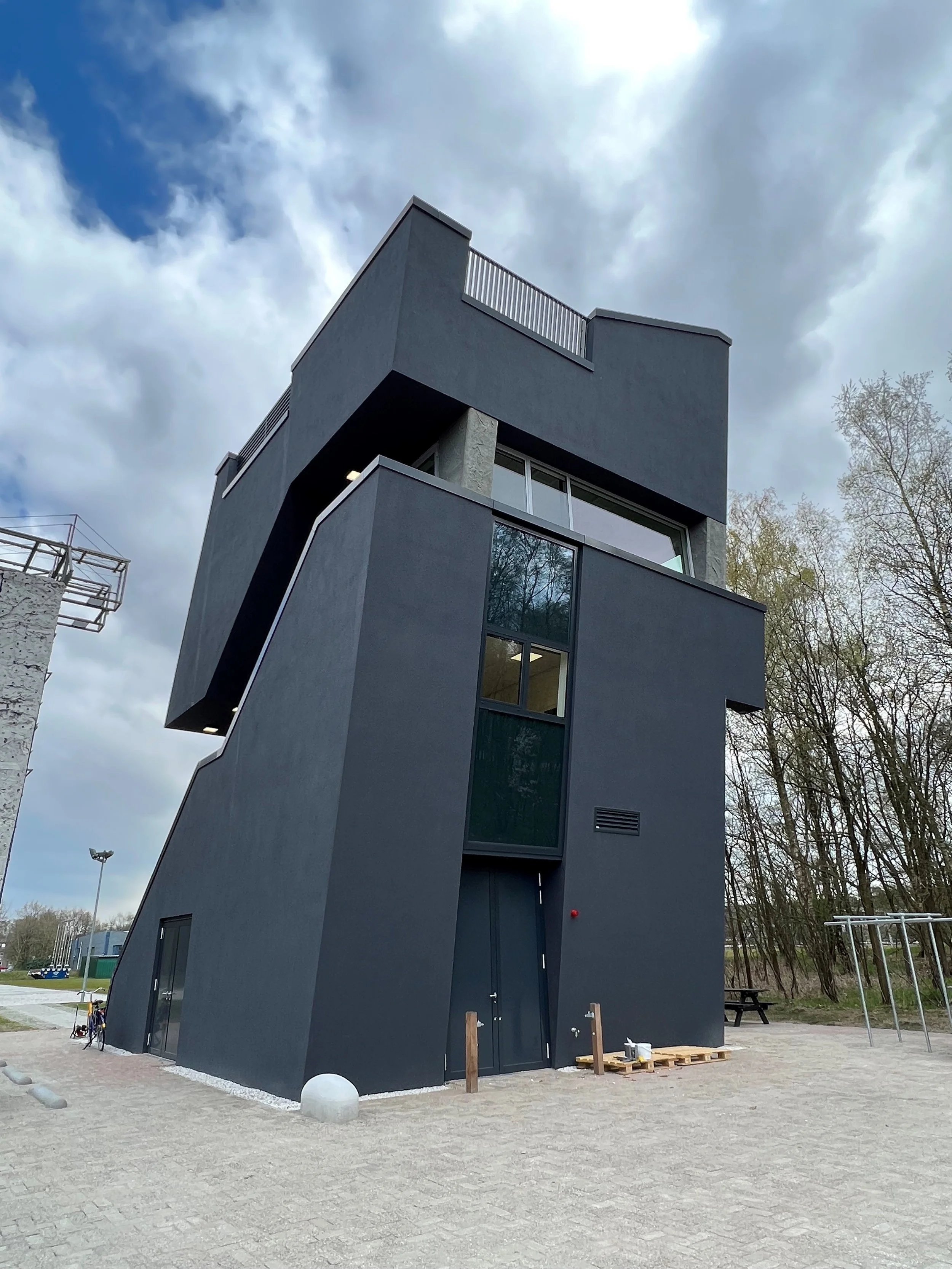 Moderne zwarte driedelige woning met grote ramen en een balkon, omgeven door een stenen verharde tuin en bomen, onder een bewolkte hemel.