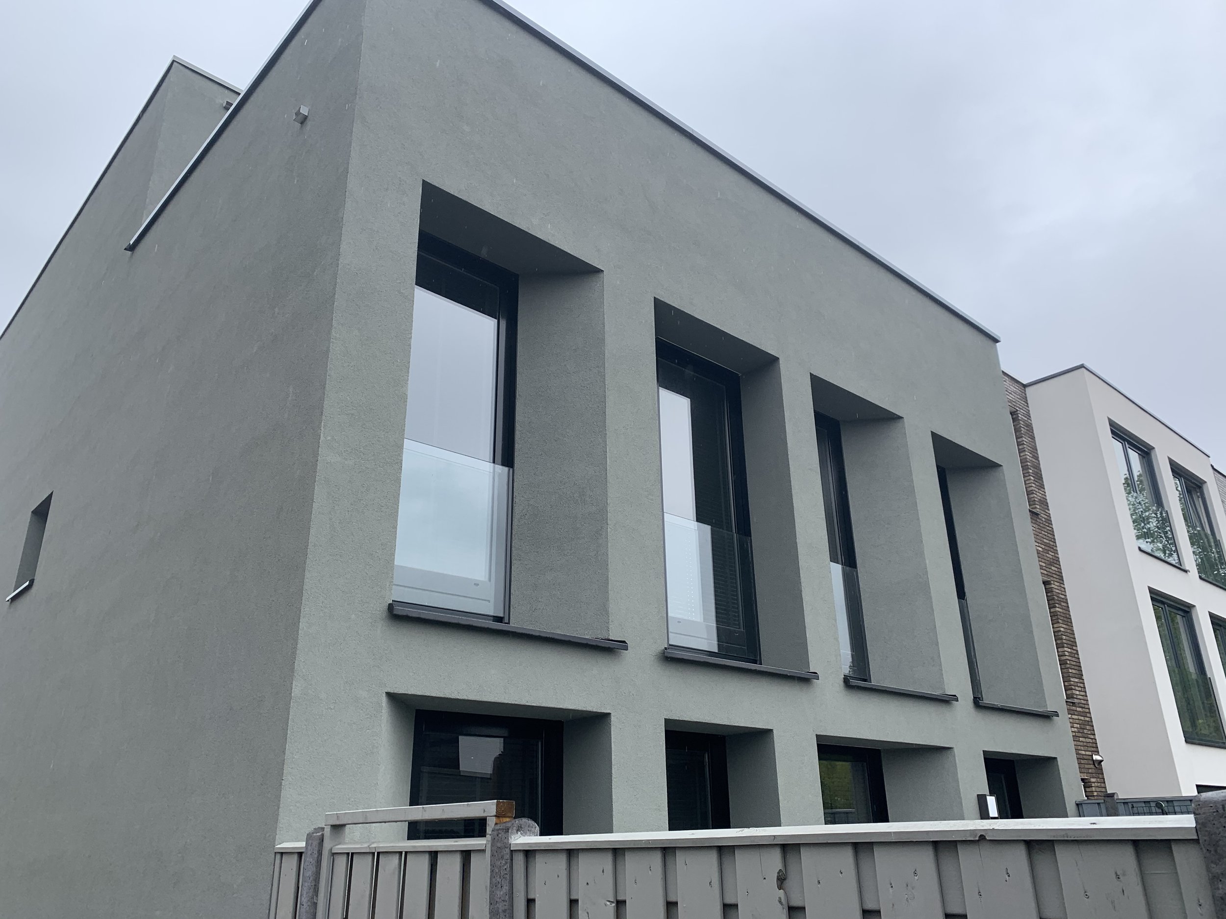 Moderne, groene appartementsgebouw met grote ramen en balkonramen, deels onder een overhang, met een hek voor.