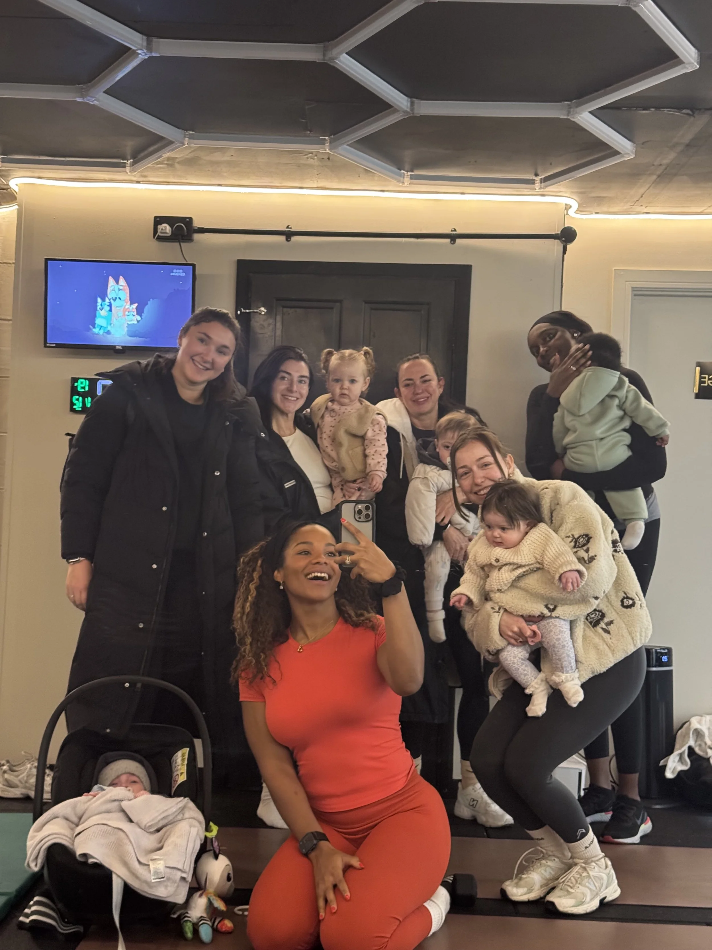 Group of women and children smiling for a photo in a gym or fitness studio, with a baby in a stroller in the foreground and some toys on the floor.