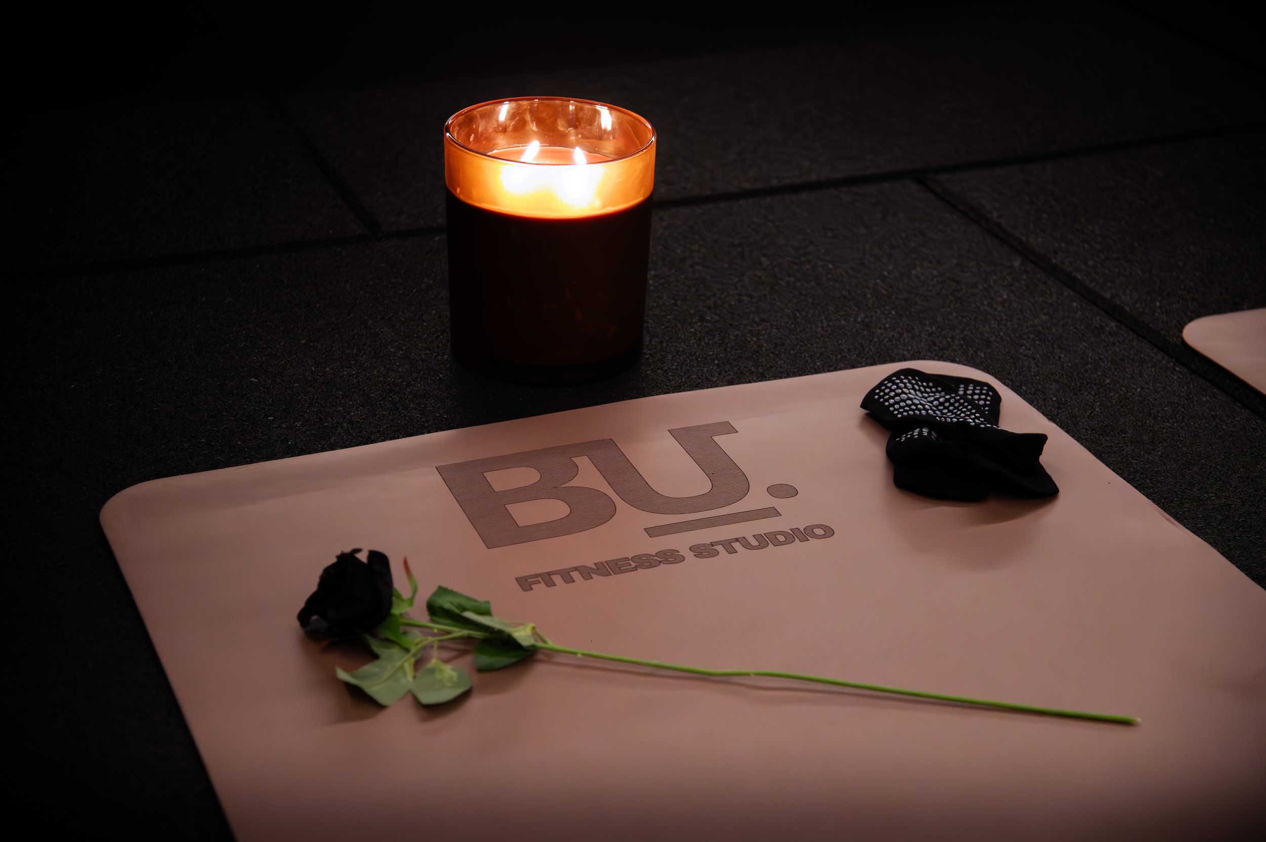 A yoga mat with the words 'BU FITNESS STUDIO', a black rose, black workout gloves, and a lit candle on a black tiled floor.