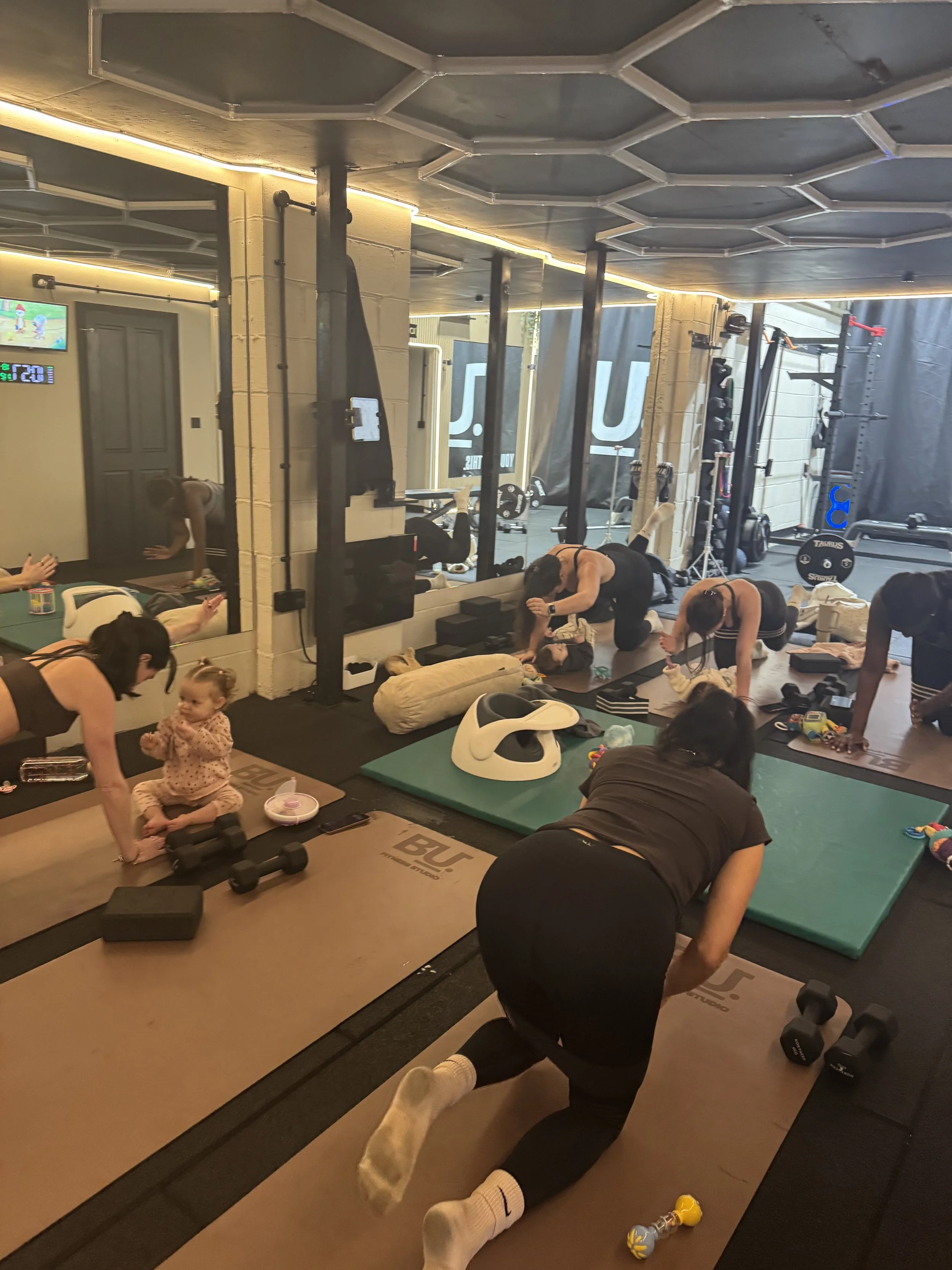 A group of women and a child participating in a workout class at a gym, using exercise mats and equipment, with some women doing planks and others doing different exercises.