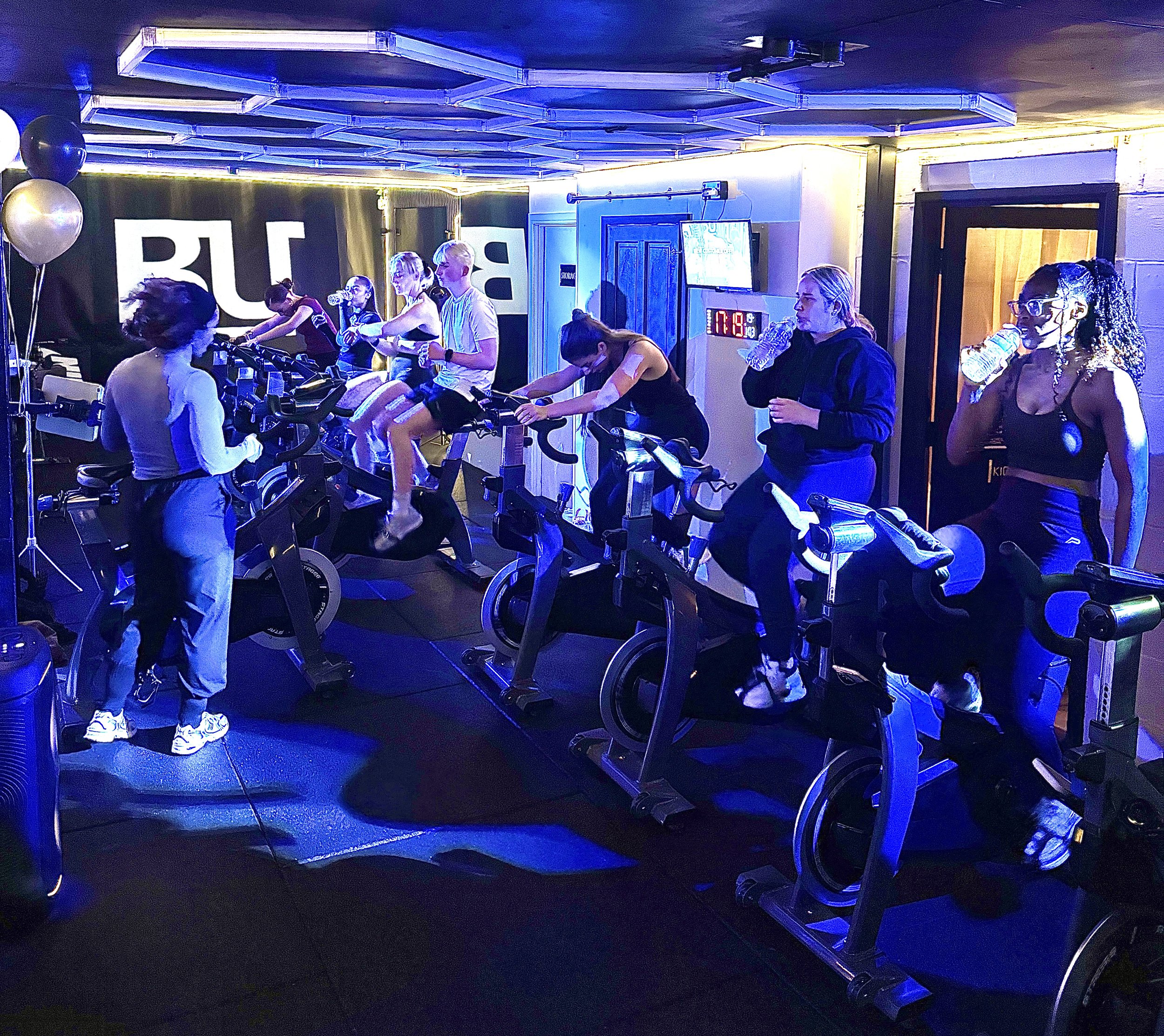 People participating in a group cycling class in a dark room with blue lighting, several individuals on stationary bikes, some drinking water, a person instructing, and a large illuminated sign with the letters 'BU' in the background.