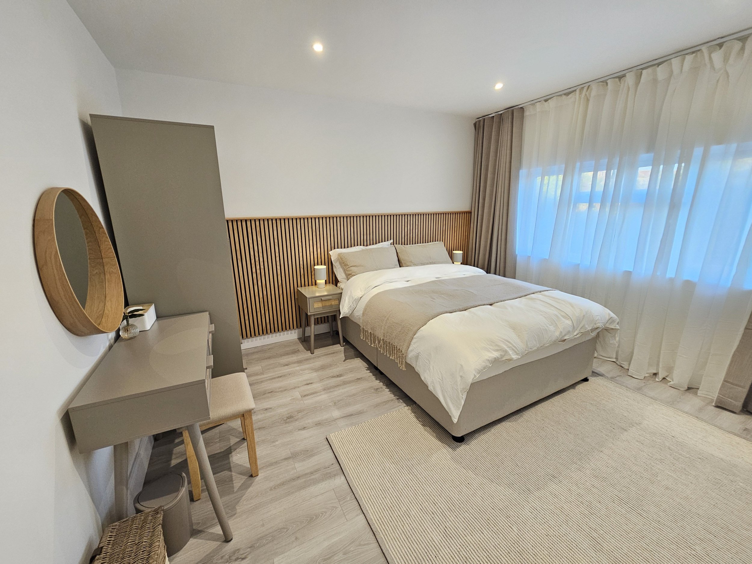 Modern bedroom with a large bed, bedside tables with lamps, a vanity with a mirror, a desk with a stool, and large windows with sheer curtains, in a neutral color palette.