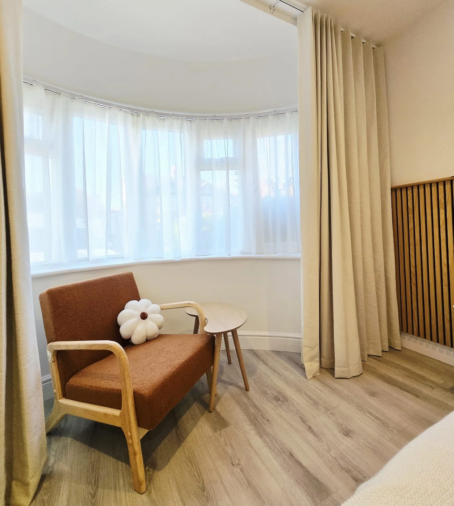 A cozy room with a brown armchair, a small side table, and a flower-shaped cushion, near a large curved window with sheer white curtains and beige drapes.편안한 의자, 넓은 채광, 런던, 한인민박,하나스테이