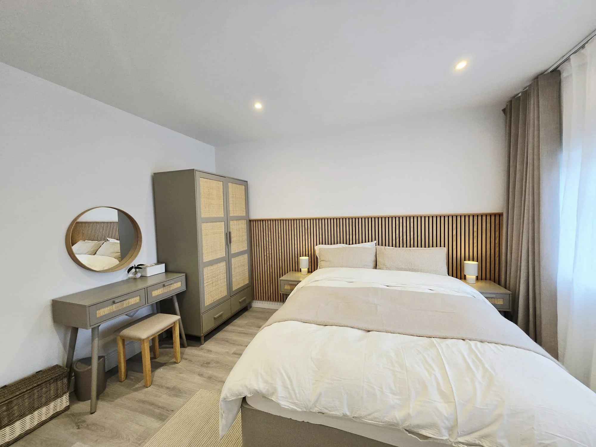 Modern bedroom with a wooden headboard, neutral bedding, nightstands with lamps, a wardrobe, a vanity with a mirror, curtains, and light-colored flooring.