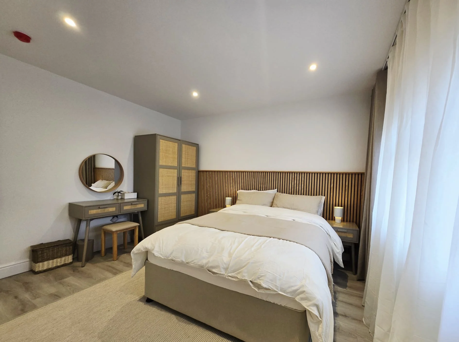 A modern bedroom with a large bed, white bedding, wooden headboard, nightstands with lamps, a wardrobe, a vanity with a mirror, a stool, and a basket, with natural light coming through curtains.