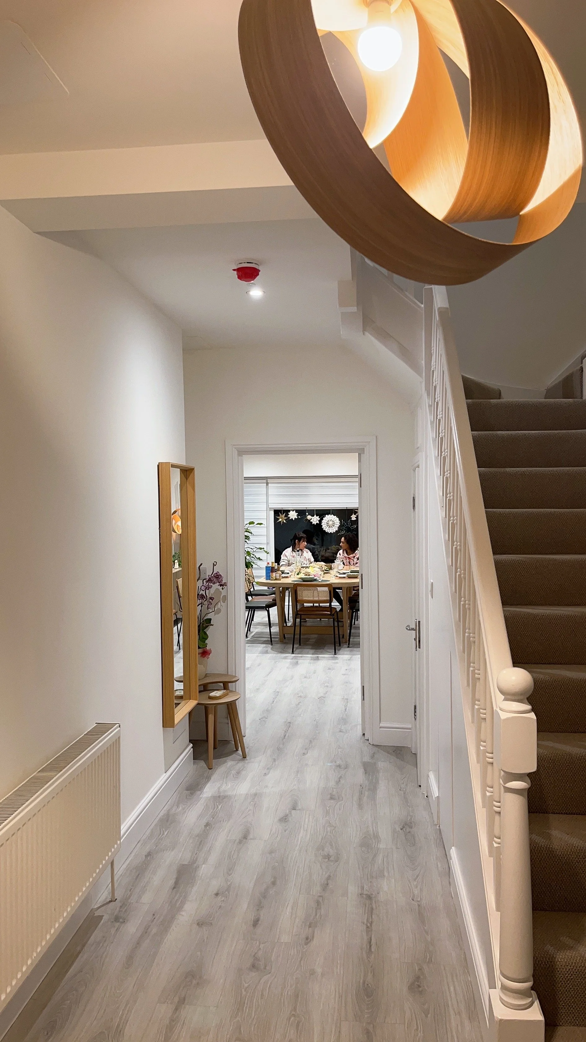 View of a hallway leading to a dining room with a table and people, seen from the living area with stairs and a modern hanging light fixture.아늑한 홀웨이, 멋진 조명, 런던, 한인민박, 하나스테이