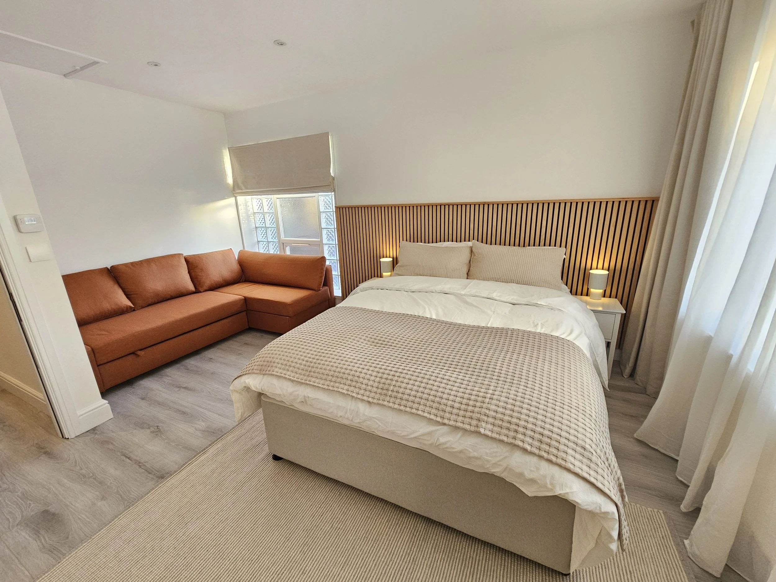 A bedroom with a double bed and two bedside tables with lamps, a brown sofa, light-colored walls, and wooden flooring.
