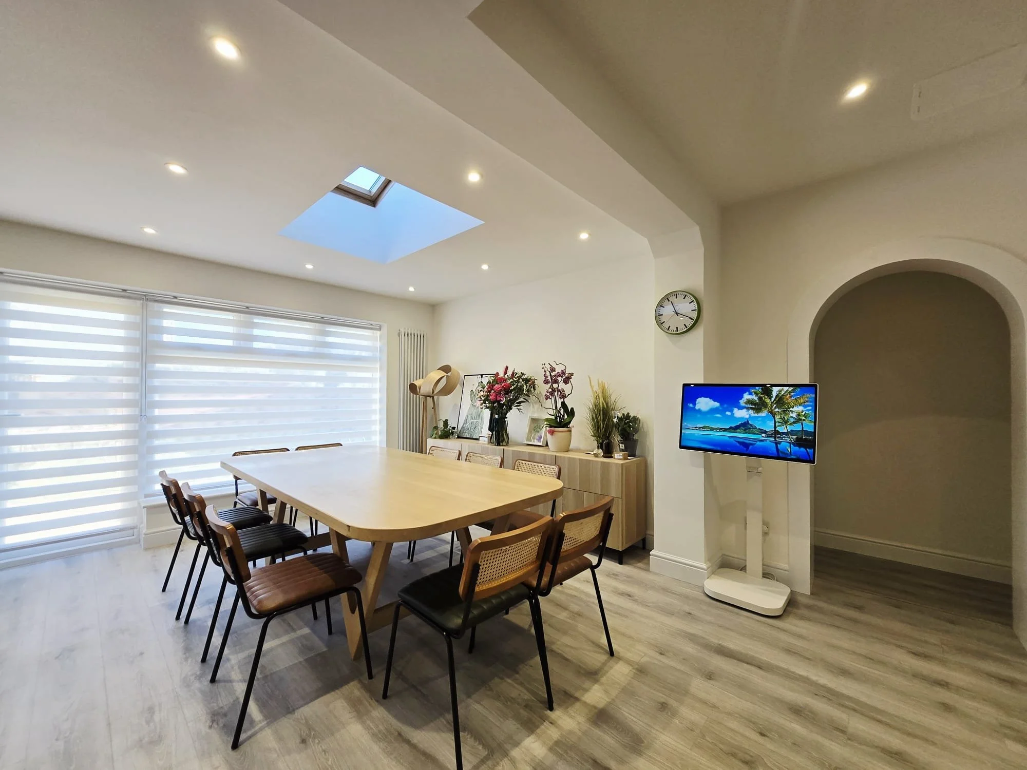 Bright dining room with a large wooden table, eight chairs, a sideboard with potted plants and flowers, a wall clock, a TV displaying a tropical beach scene, and a skylight in the ceiling.