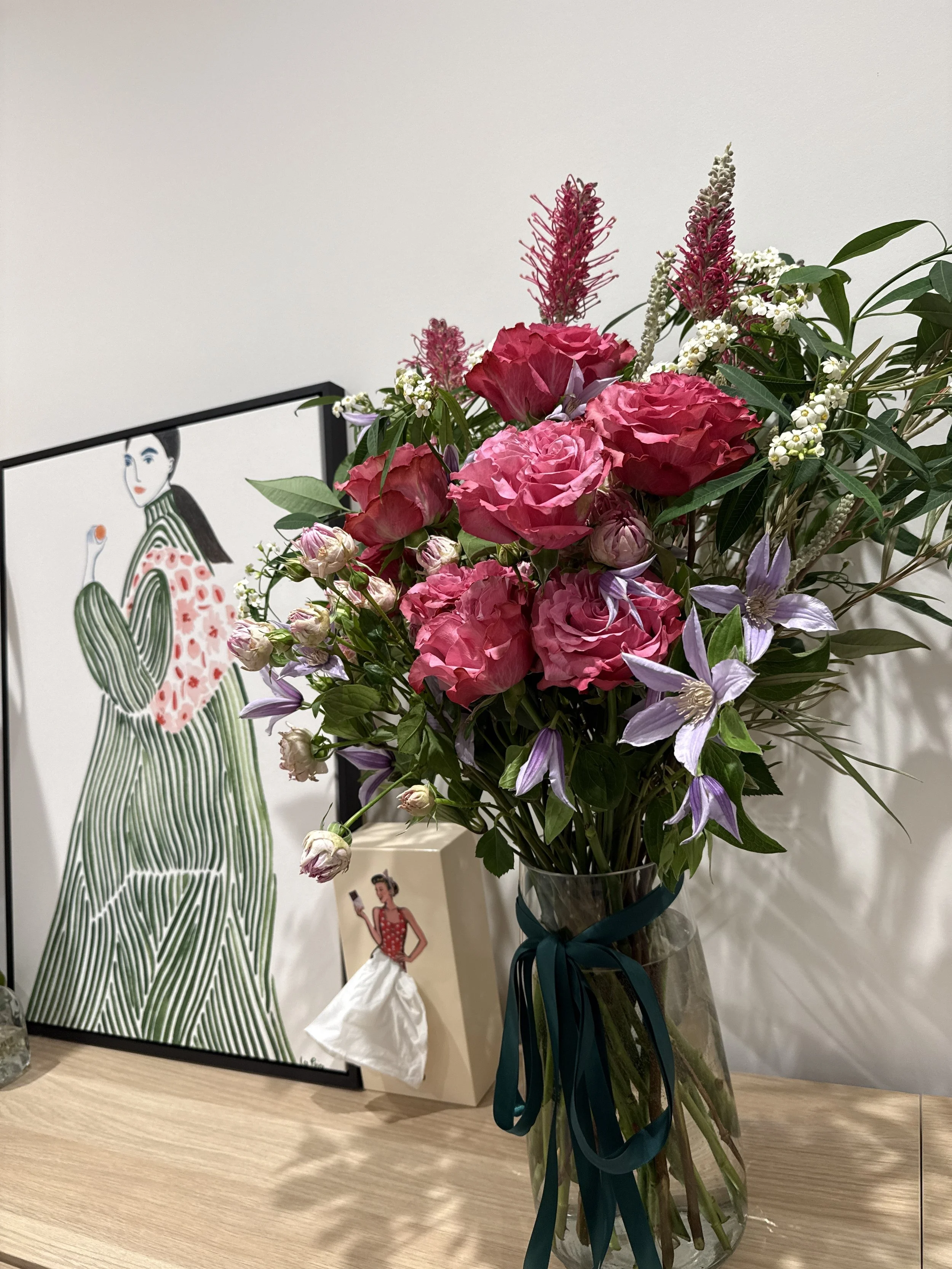 A bouquet of pink and purple flowers in a glass vase with a teal ribbon, placed on a wooden surface against a white wall, with artwork of a woman in a striped green and white dress in the background.