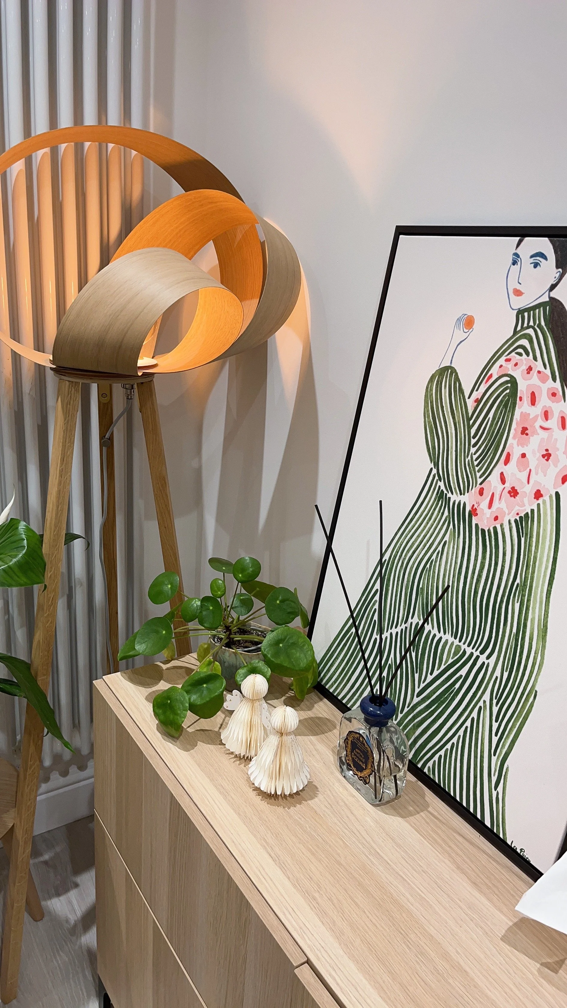 Decorative interior scene with a wooden sideboard, green potted plant, franchising figurines, a framed illustration of a woman in green and pink floral clothing, a modern lamp, and a diffuser.런던,한인민박, 감각적 인테리어 조명