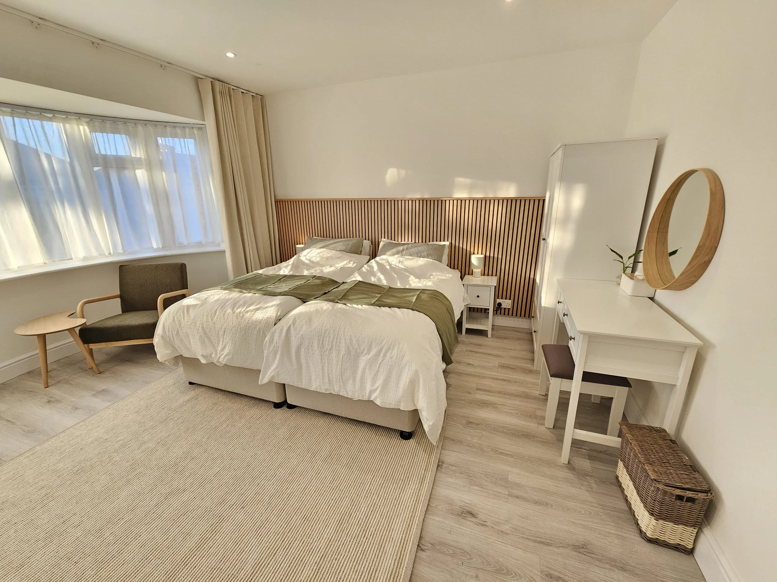 A cozy bedroom with two beds pushed together, a wooden headboard, a nightstand with a lamp, a chair near the window, a white dresser with a mirror and plant, and a woven basket on the floor, illuminated by natural light from large windows with curtains.