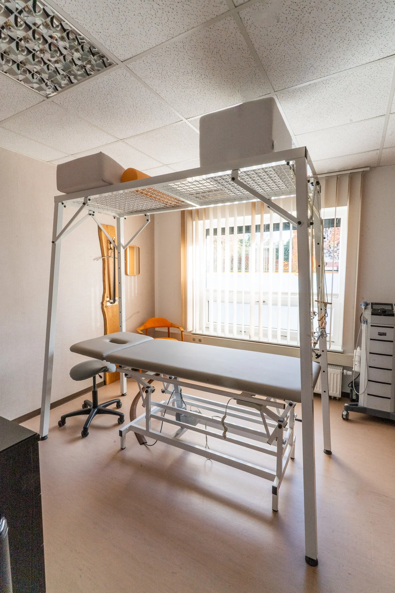 Medical examination room with an adjustable examination table, a stool, a chair, and medical equipment, natural light coming through window with blinds.