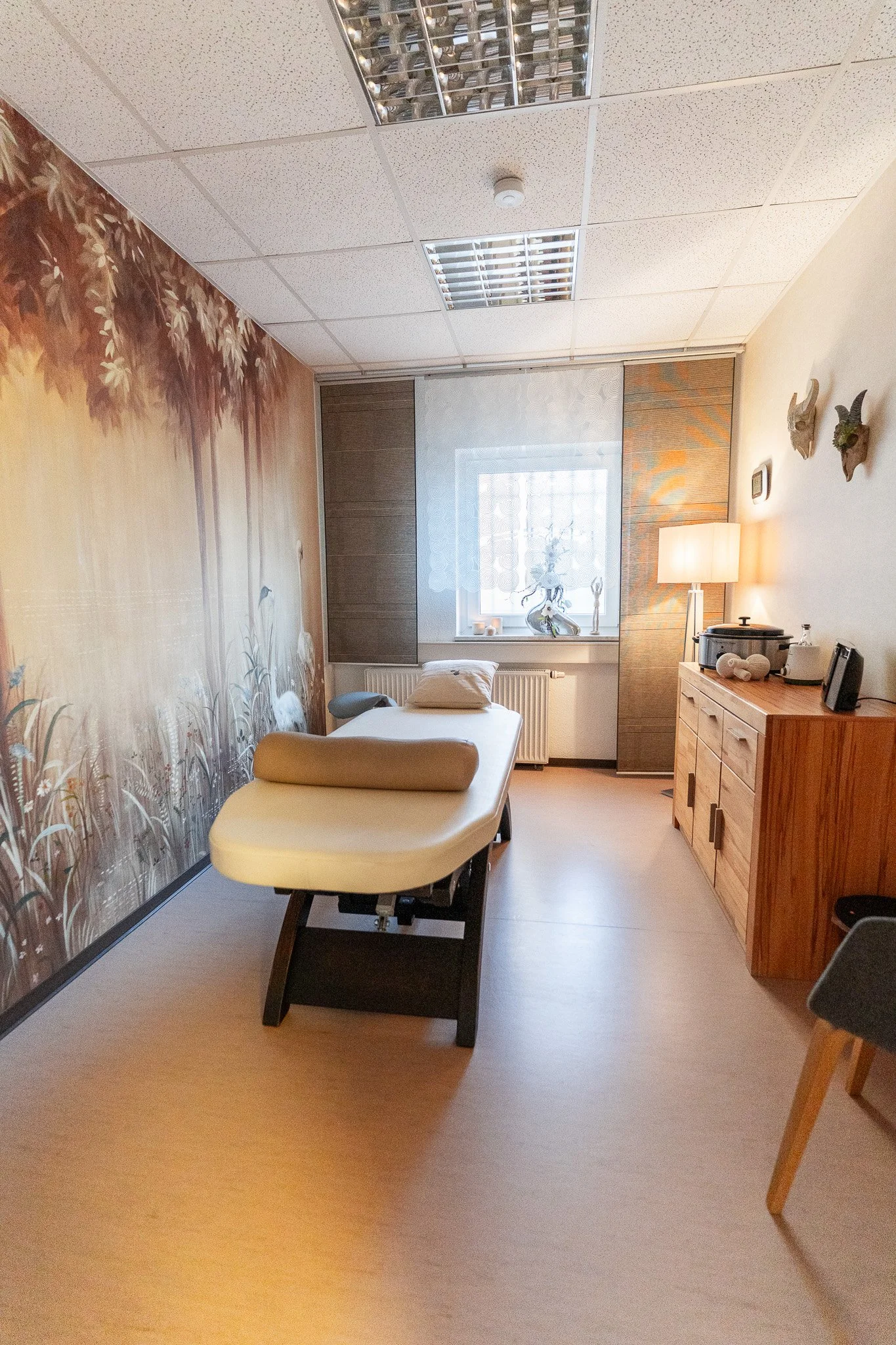 A wellness or massage therapy room with a massage table, a decorative wall mural of a forest scene, a window with sheer curtains, a wooden cabinet with various items, and warm lighting.