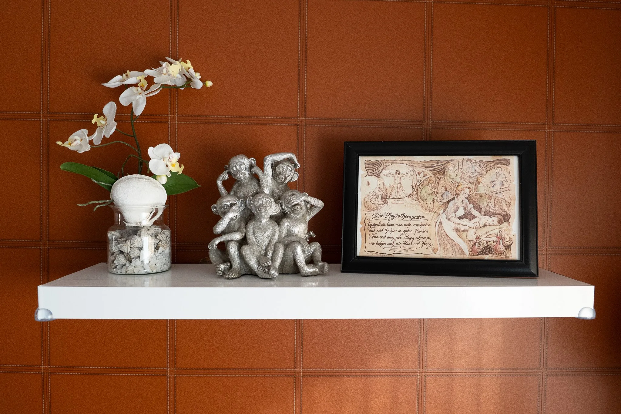 A white floating shelf on a brown-tiled wall, with a glass vase holding an orchid plant, a group of silver monkey figurines, and a framed artwork with German text and sketches.