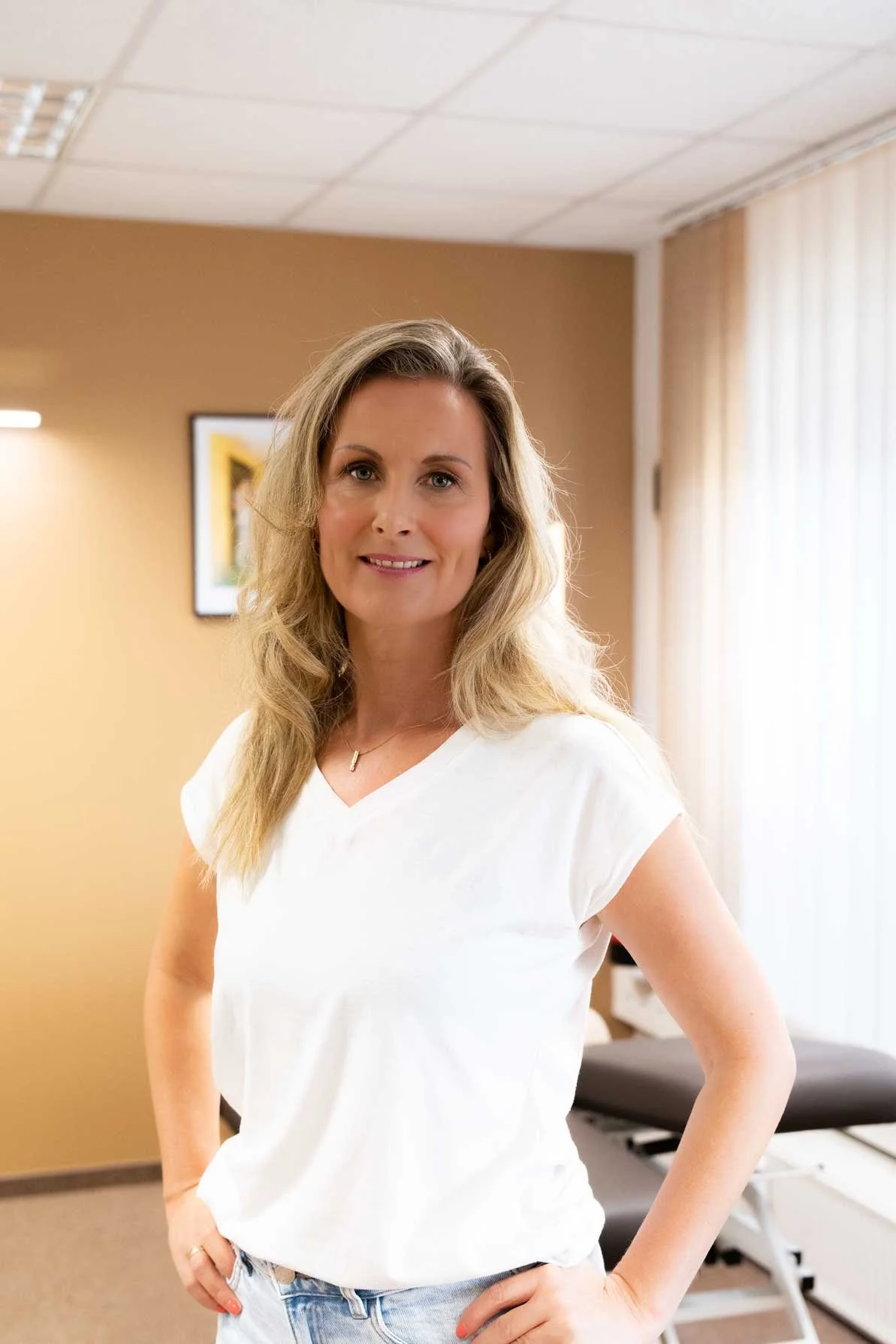 A woman with blonde hair wearing a white shirt standing in a room with medical examination table and beige walls.