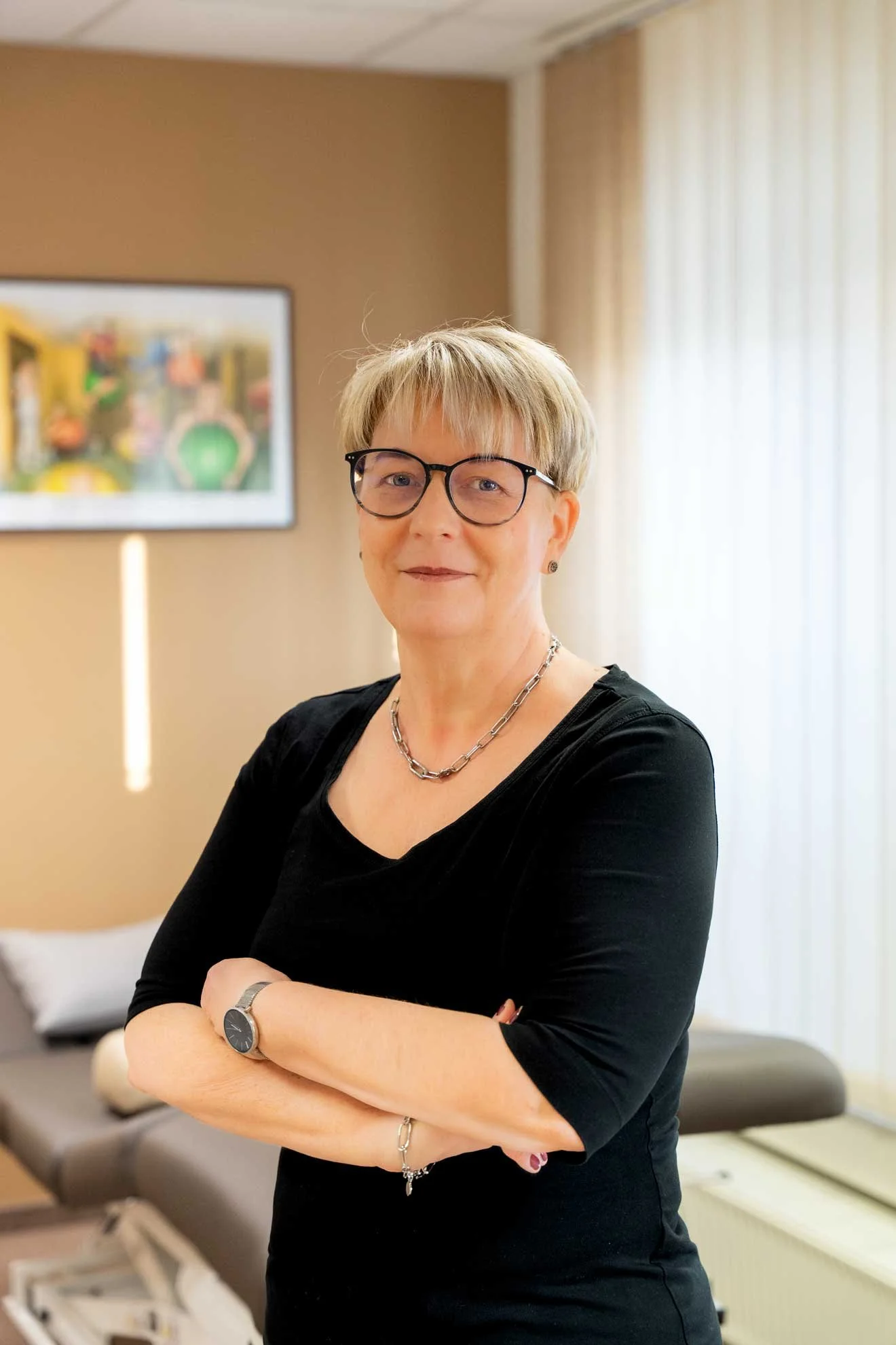 A woman with short blonde hair and glasses, wearing a black top, standing with arms crossed in a room with artwork on the wall and curtains covering a window.