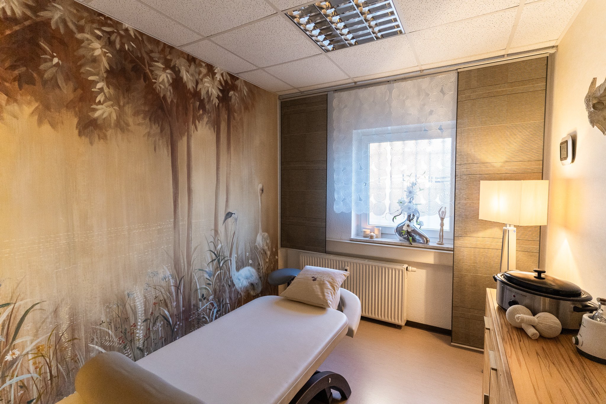 A treatment room with a massage table, wall mural of trees and cranes, window with lace curtain, and a dresser with a lamp and spa equipment.