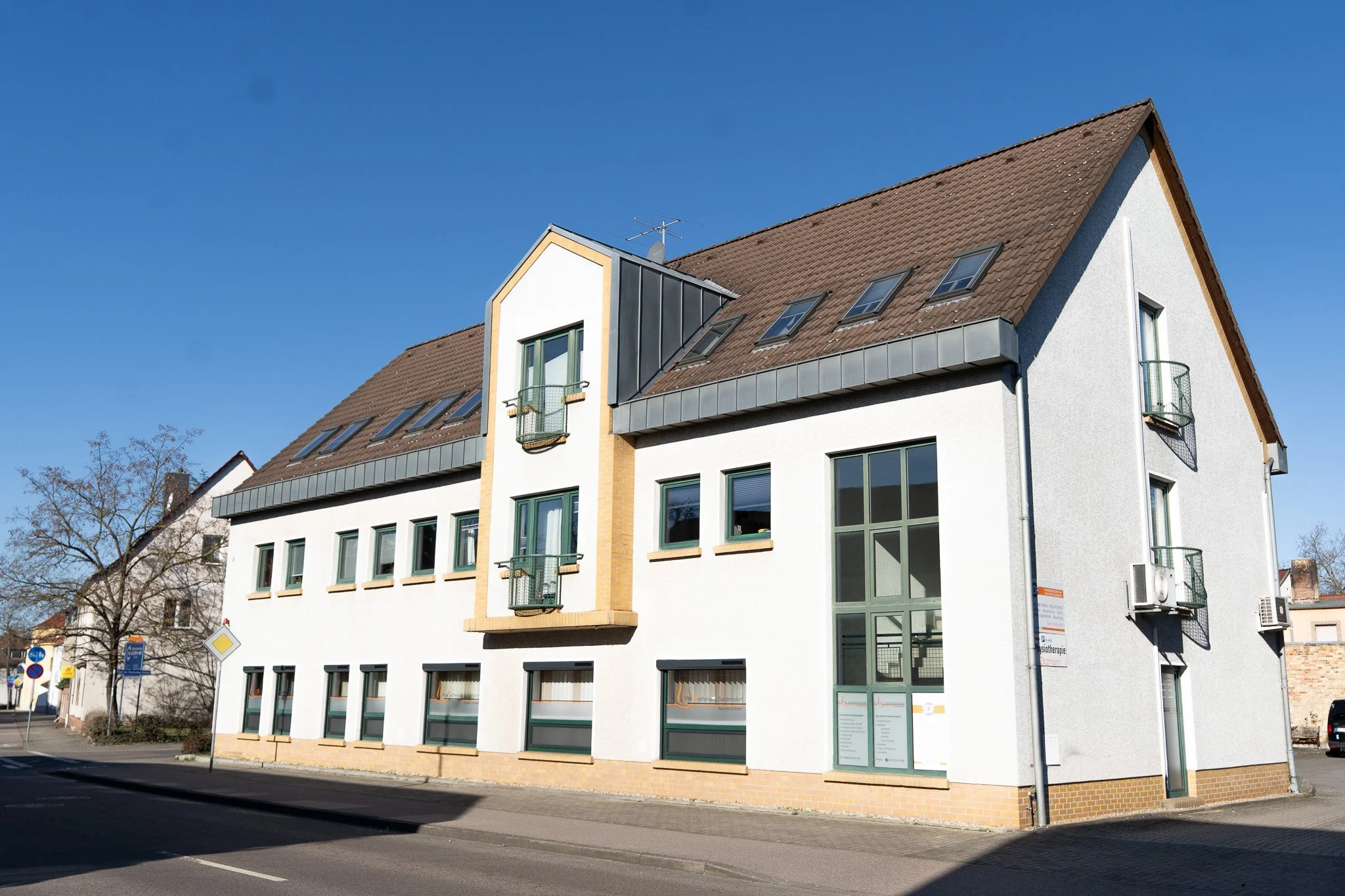 A white multi-story building with a brown gabled roof and small balconies on each side, large windows, and a glass entrance with signs, located along a street under a clear blue sky.