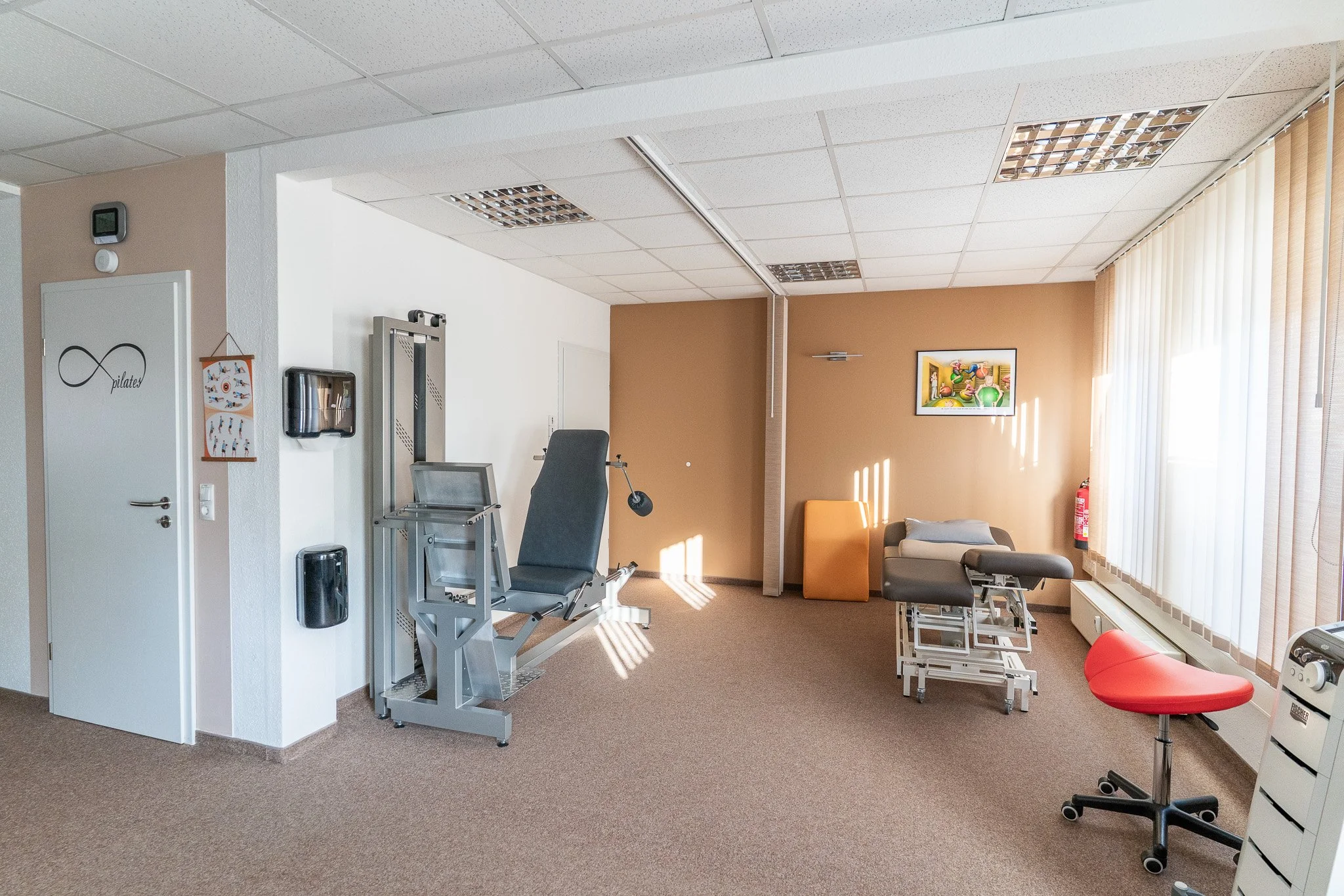 A physical therapy or rehabilitation room with specialized equipment, including an adjustable treatment bed, an exercise machine, a red stool, and a therapy table, with natural light coming through vertical blinds and artwork on the wall.