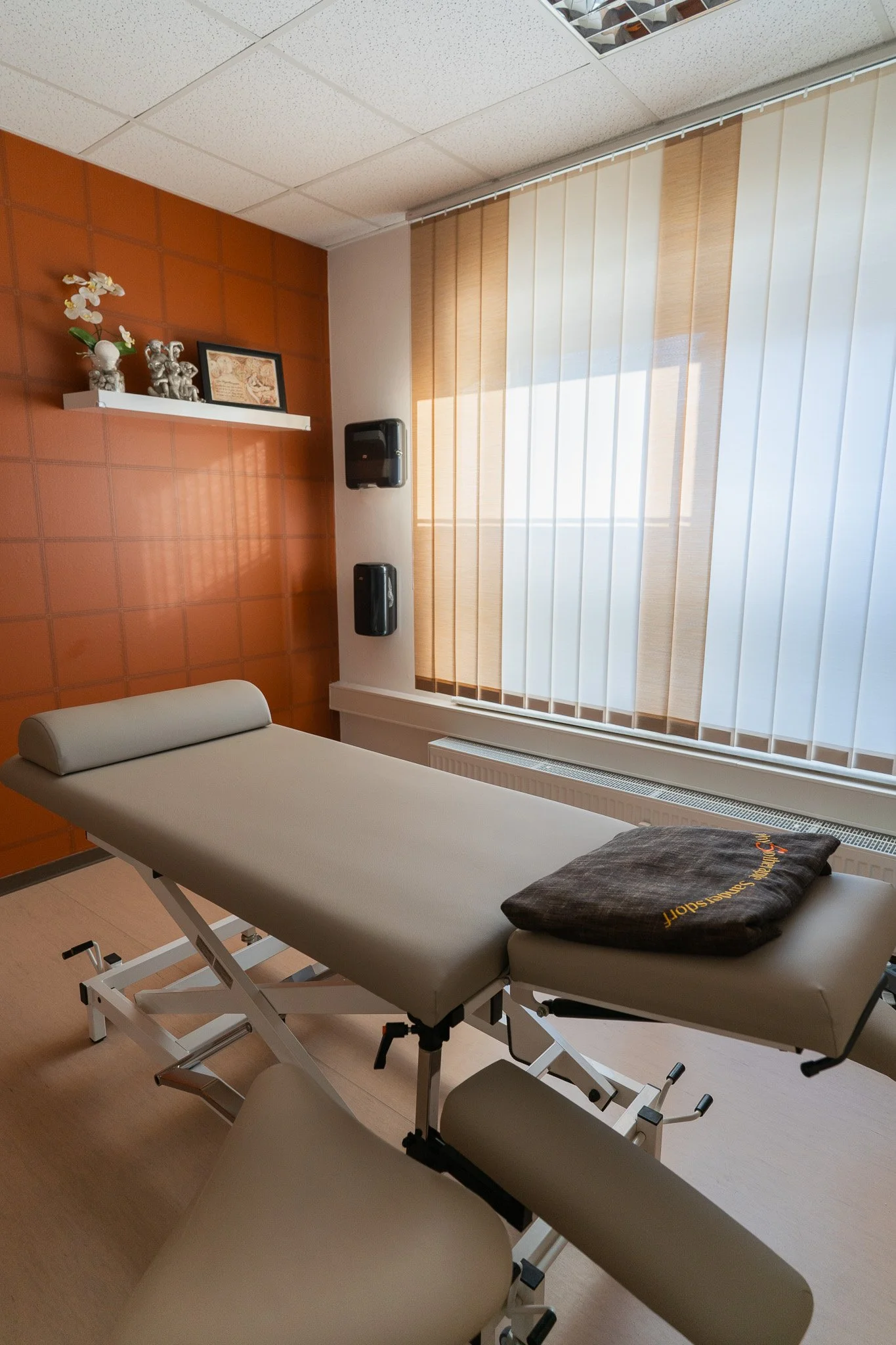 Treatment room with an adjustable massage or examination table, a window with vertical blinds, and decorative items on a wall shelf.