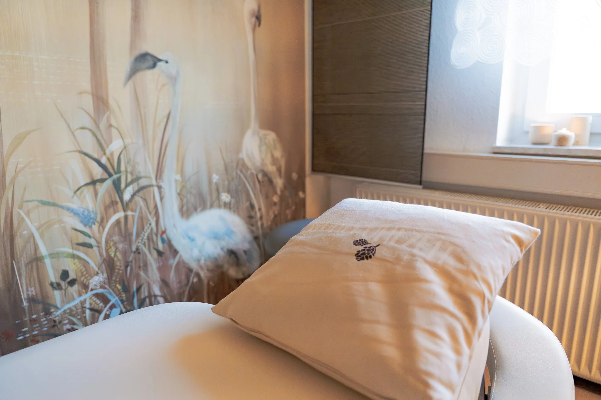 A bedroom with a white bed and a pillow with black leaf pattern, a floral wallpaper featuring cranes and grass, a window with candles, and a radiator.