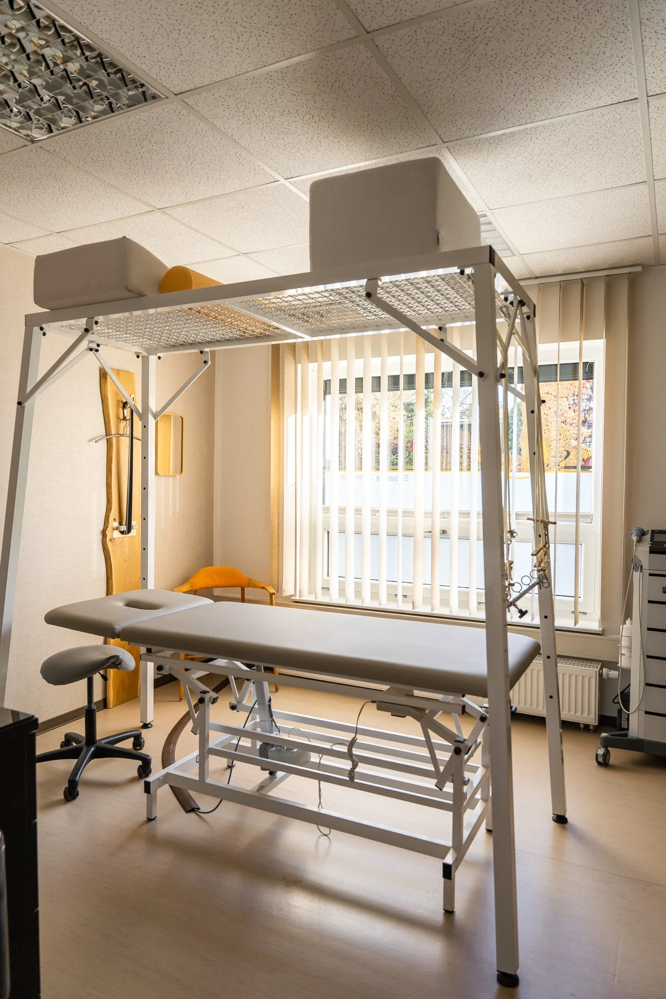 Medical examination room with a treatment table under a canopy, a rolling stool, a wooden wall hook, a small yellow chair, a window with blinds, and medical equipment.