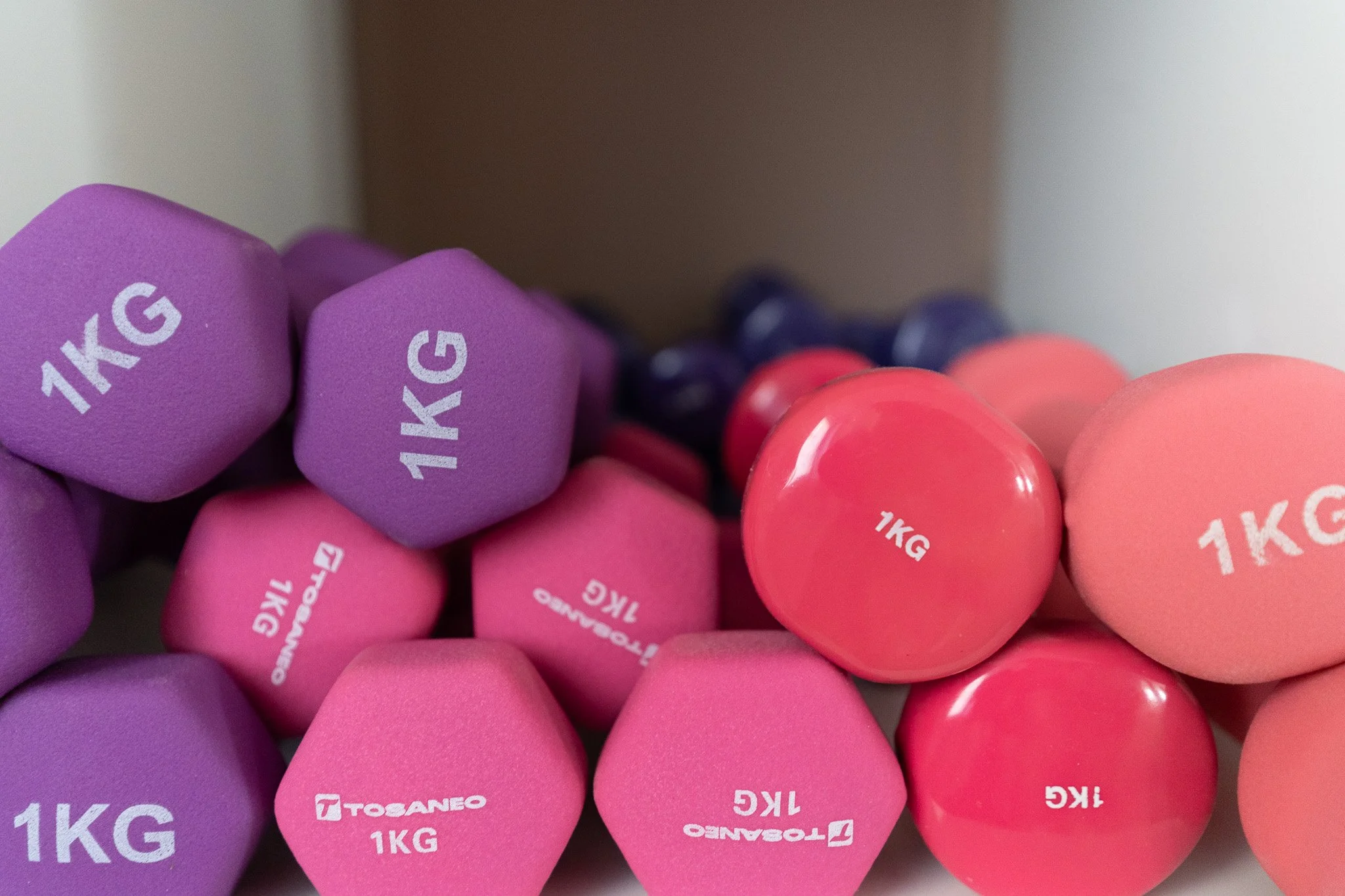 A collection of multicolored dumbbells, mainly purple and pink, with some dark blue ones in the background, all labeled '1KG'.