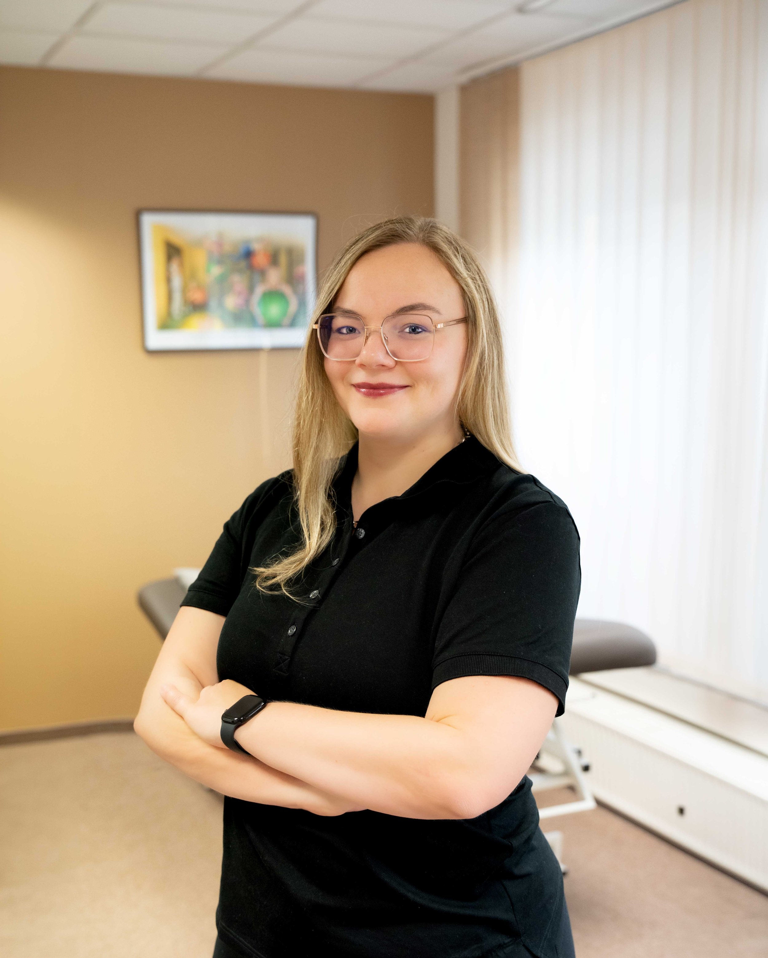 A woman with light skin and glasses standing with arms crossed in a room with beige walls, a window with blinds, and a framed colorful artwork on the wall behind her.