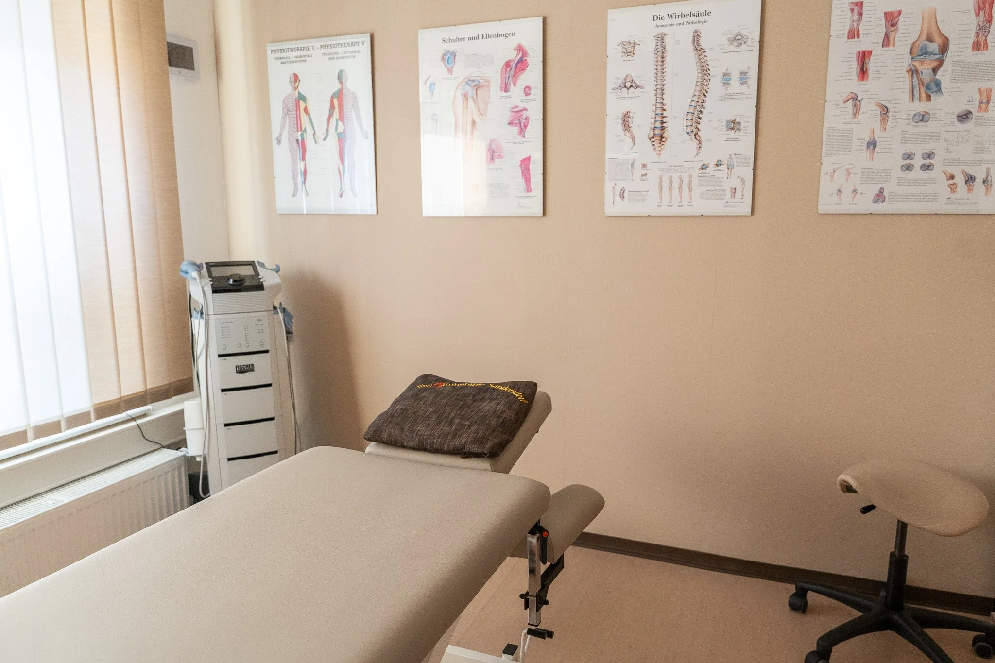 A medical examination room with a massage or therapy table, a folded brown towel on the headrest, a rolling stool, and posters of human anatomy on the wall.