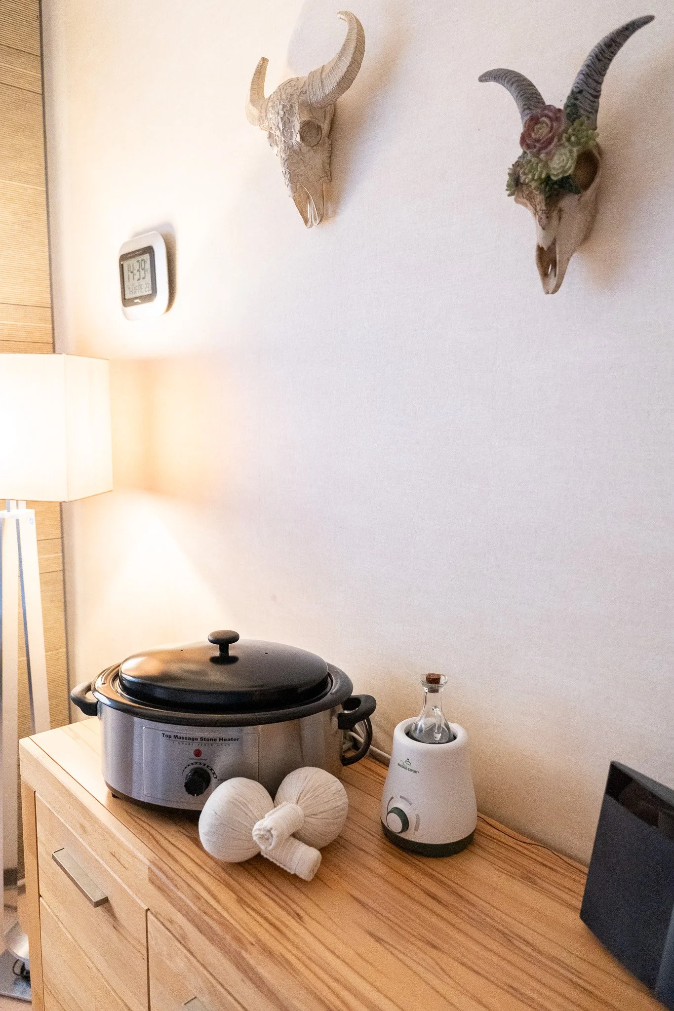 Decorative animal skulls mounted on the white wall, one with flowers and the other plain, with a digital clock, a lamp, and a slow cooker on the wooden dresser below.