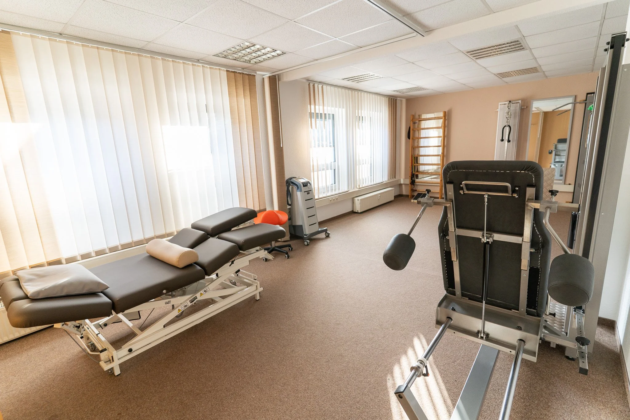 A spacious physical therapy room with a treatment table, exercise equipment including a leg press machine, and natural light coming through large windows with vertical blinds.