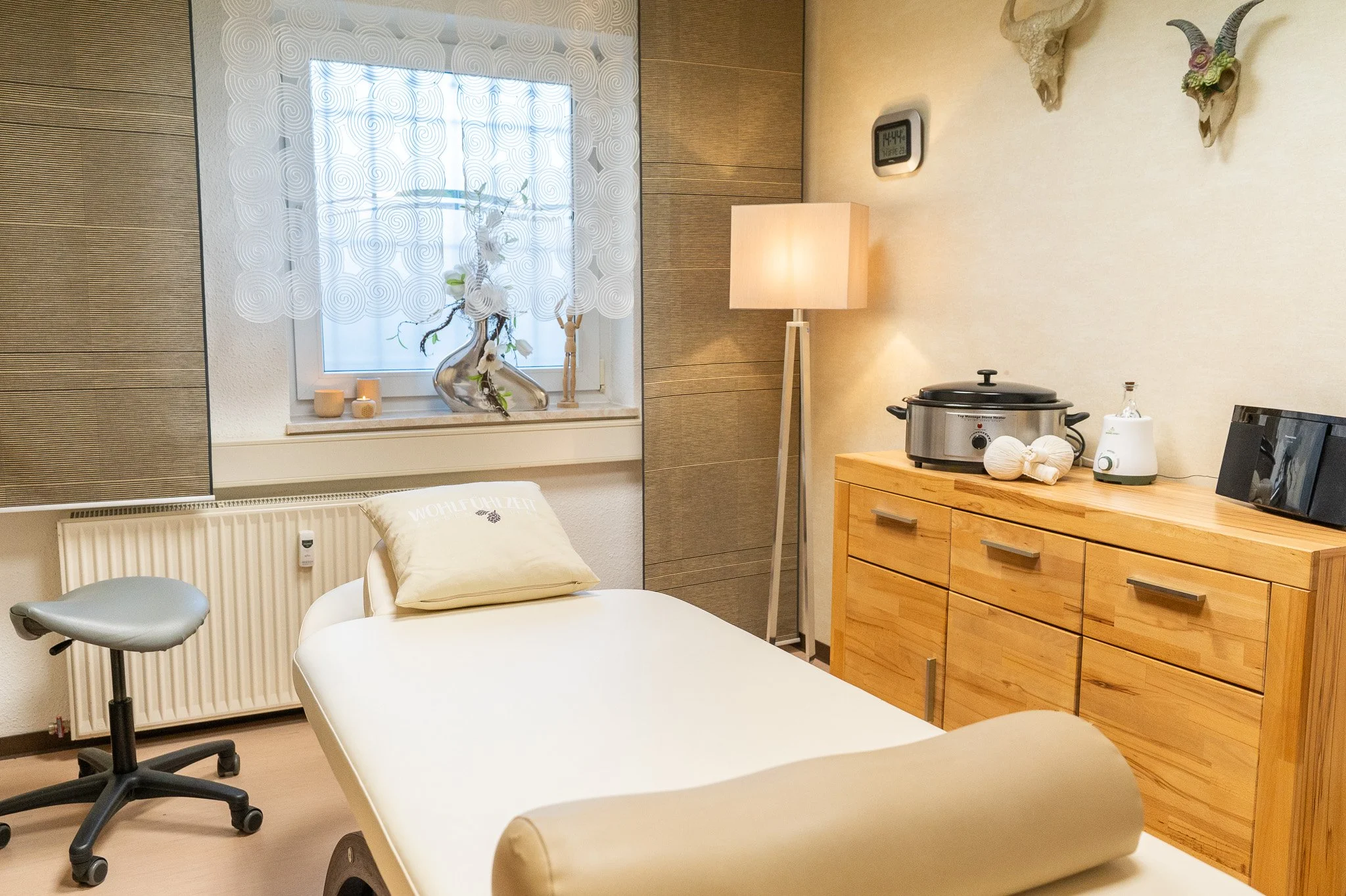 Medical examination room with an examination bed, a window with decorative curtains, a wooden cabinet with medical equipment, a lamp, and wall decorations.