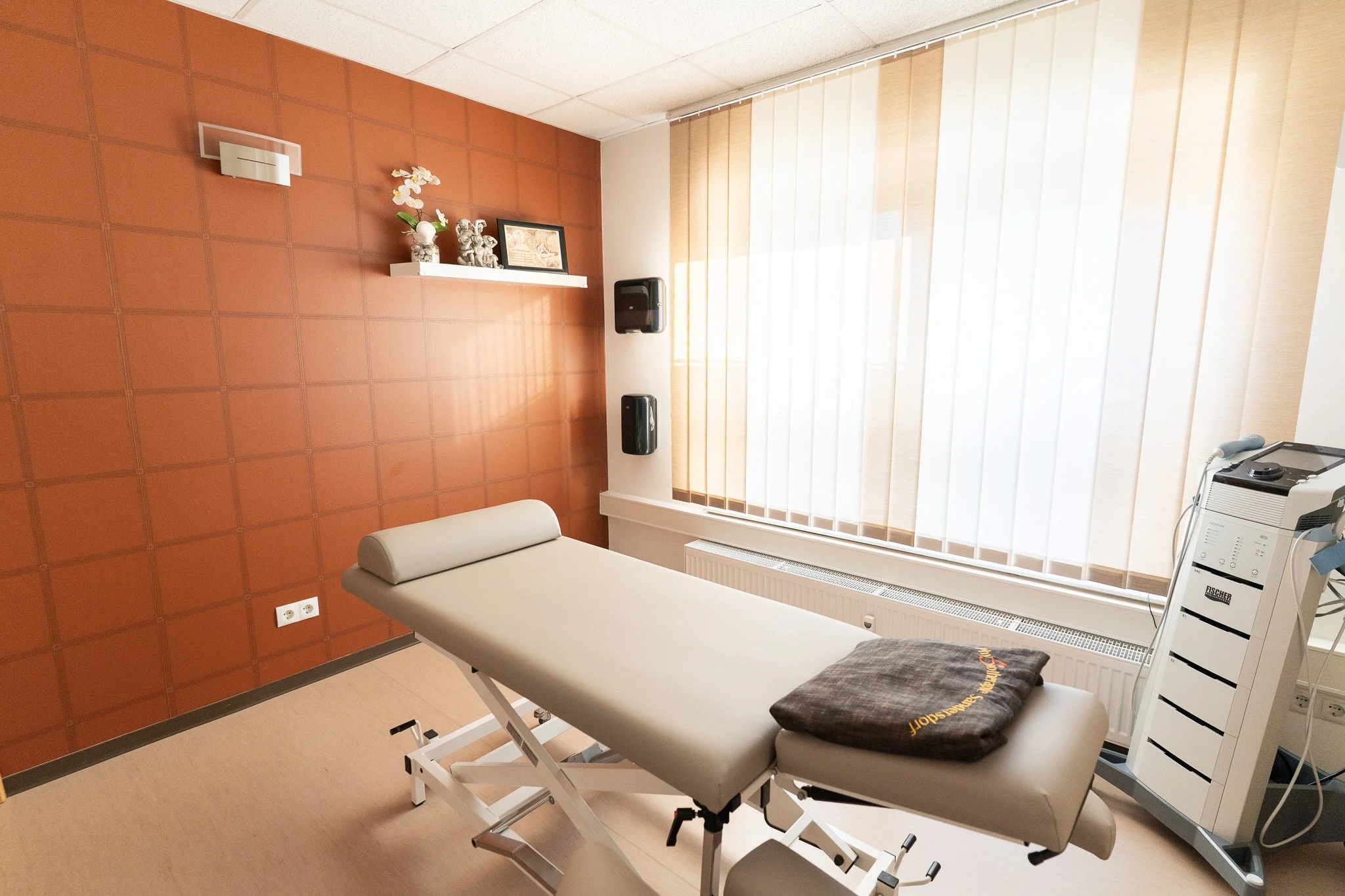 Medical examination room with a treatment bed, window with vertical blinds, a white shelf with decorative items, and medical equipment.