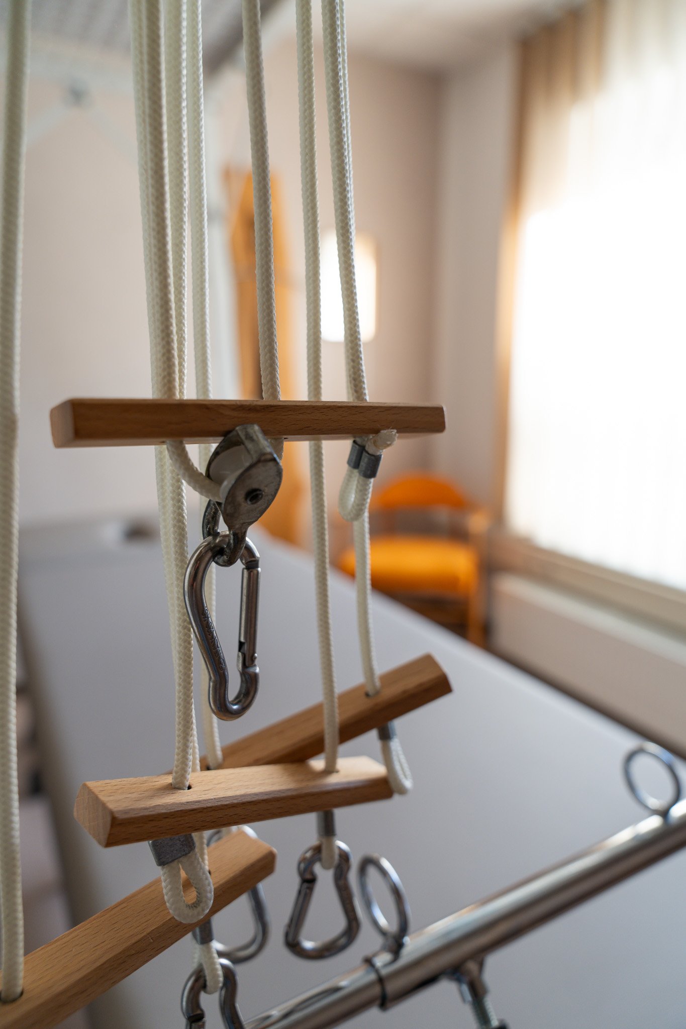An indoor playground swing set with wooden steps, ropes, metal hooks, and clips, with a blurred background of a room with natural light.