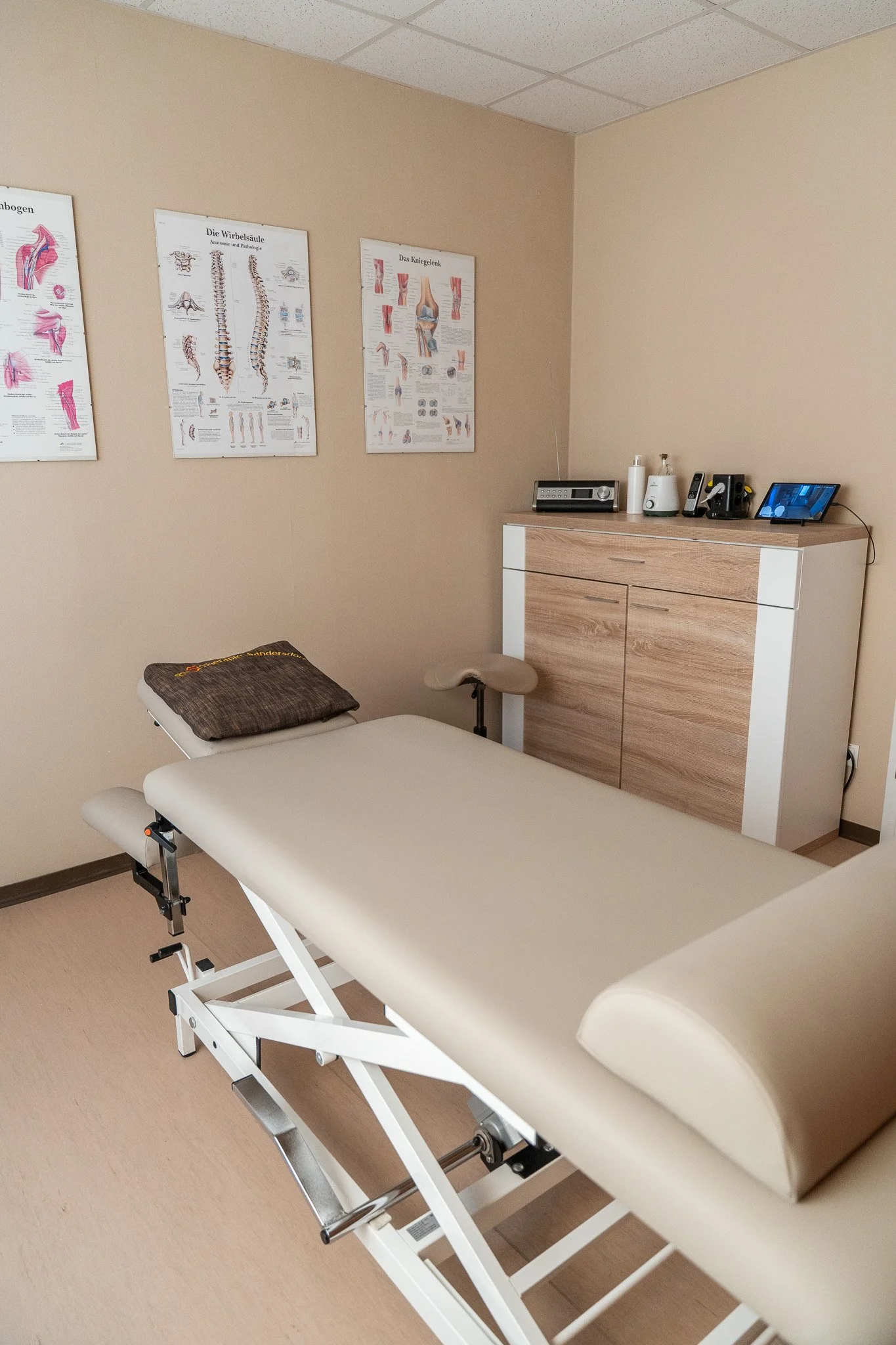 A clean, empty medical examination room with a beige medical bed, a brown towel, a wooden cabinet with medical supplies on top, and three anatomical posters on the beige wall.