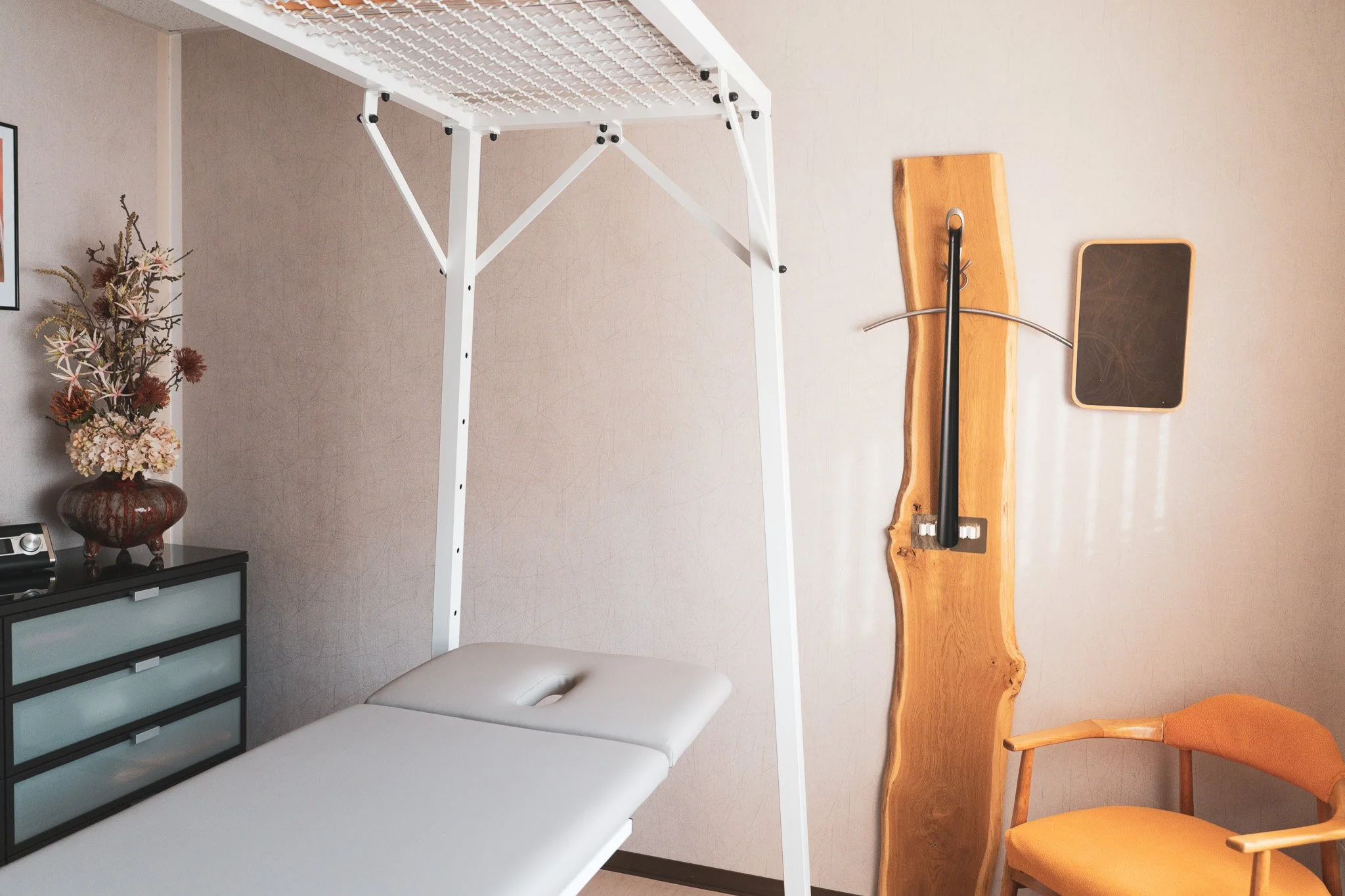 Medical examination room with a massage table, a wooden wall-mounted instrument, a small black cabinet, a vase with flowers, and wooden furniture.