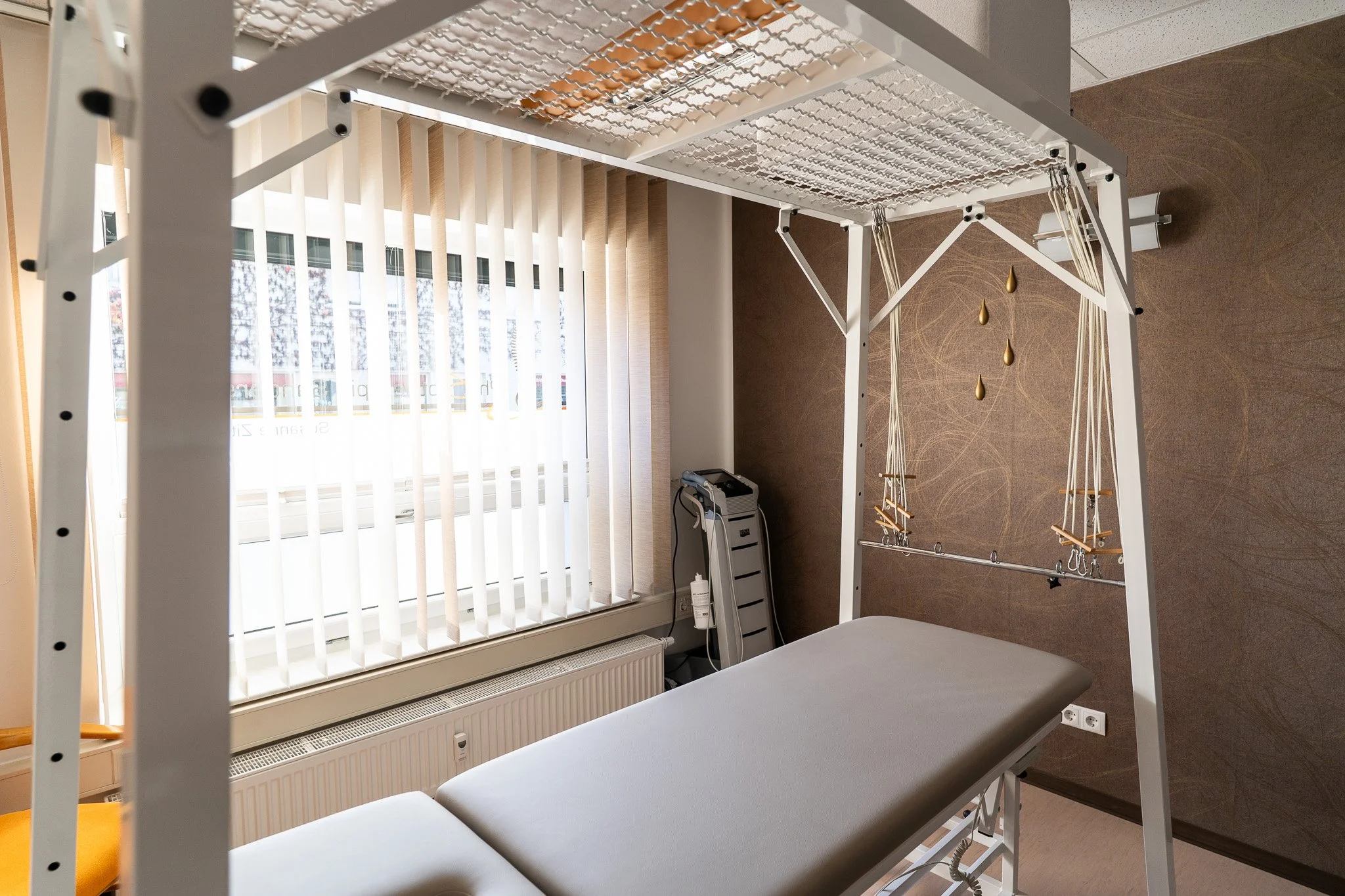 A medical examination room with a treatment table and a lightweight canopy frame with hanging straps and loops, nearby a medical device and a window with vertical blinds.