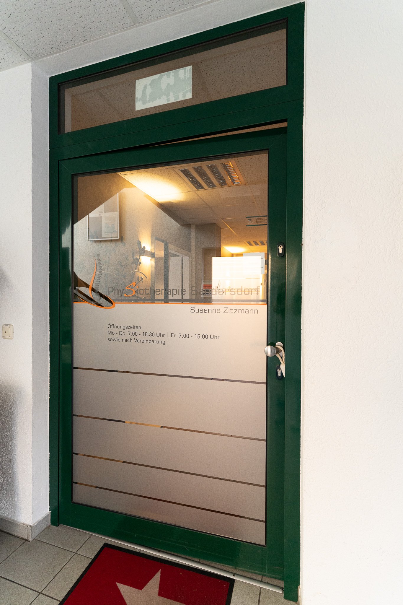 Glass door to a therapy office with green frame and frosted glass panels, displaying business hours and therapist's name in German.
