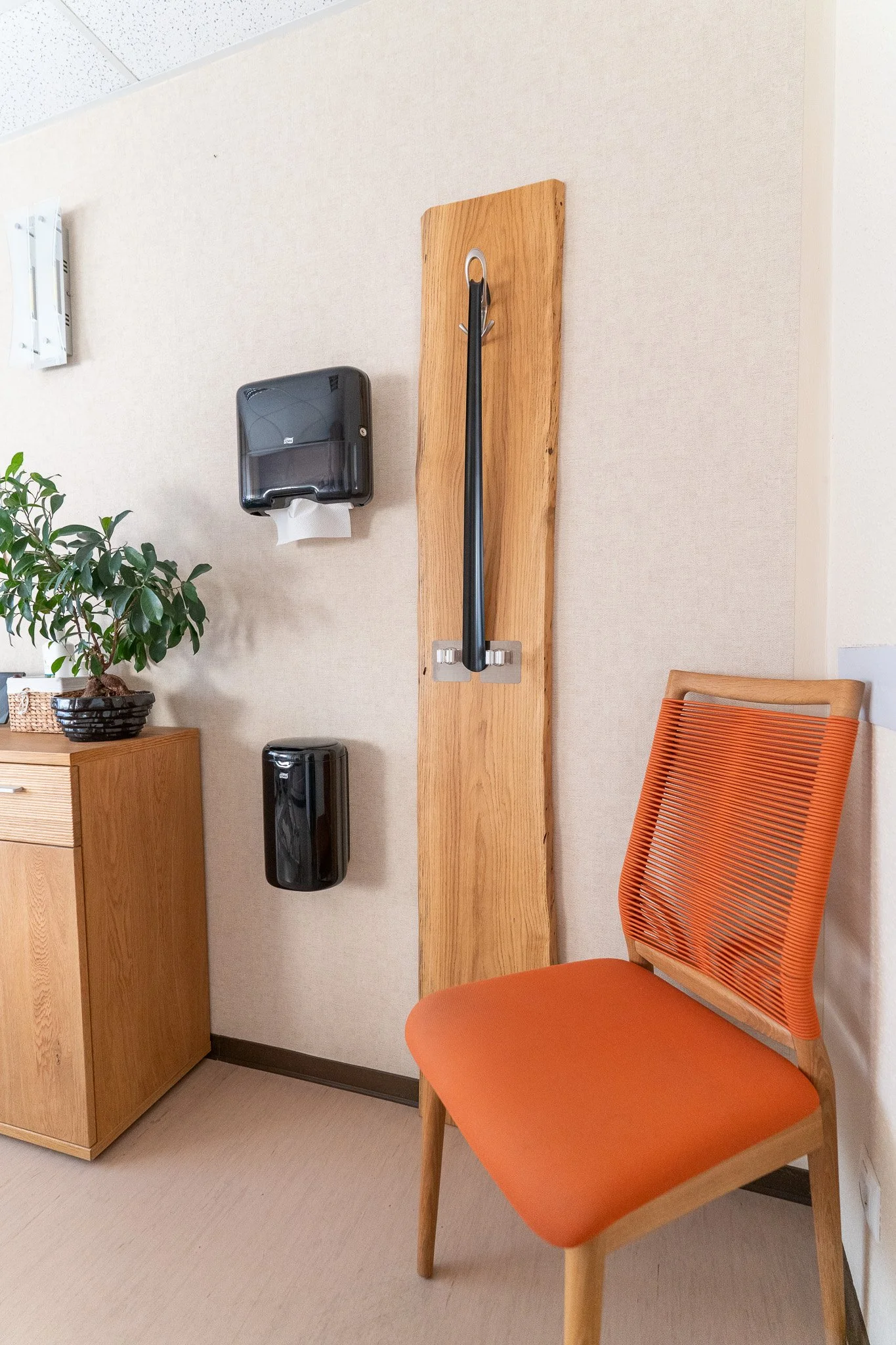 An orange chair with a wooden frame, a small wooden cabinet with a potted plant on top, and a wall-mounted wooden board with a black cane and two black dispensers in a room with beige walls and a beige floor.