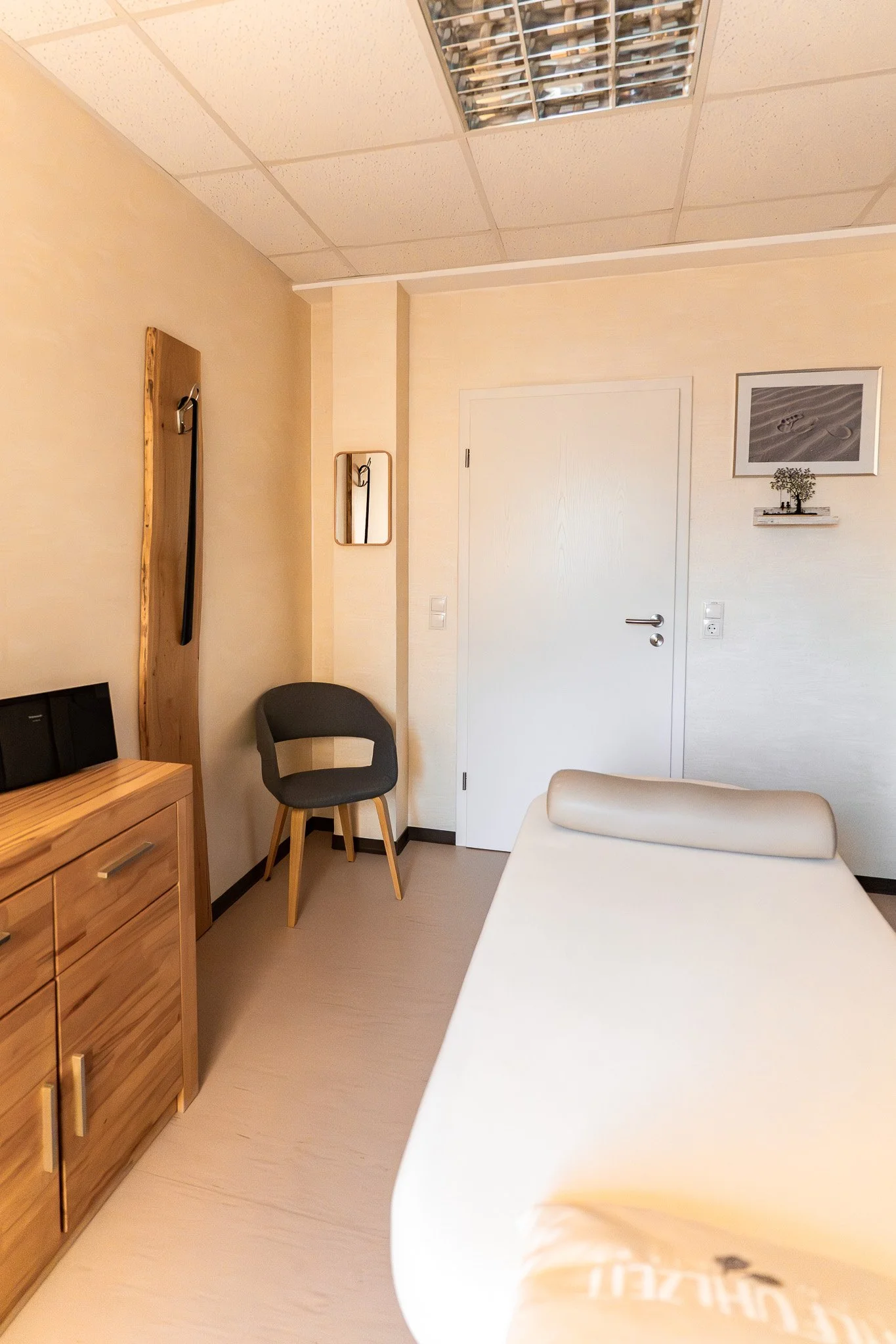 A hospital room with a bed, a wooden cabinet, a gray chair, and wall decorations, including a framed black and white photo of sand dunes and a small wooden shelf with a potted plant.