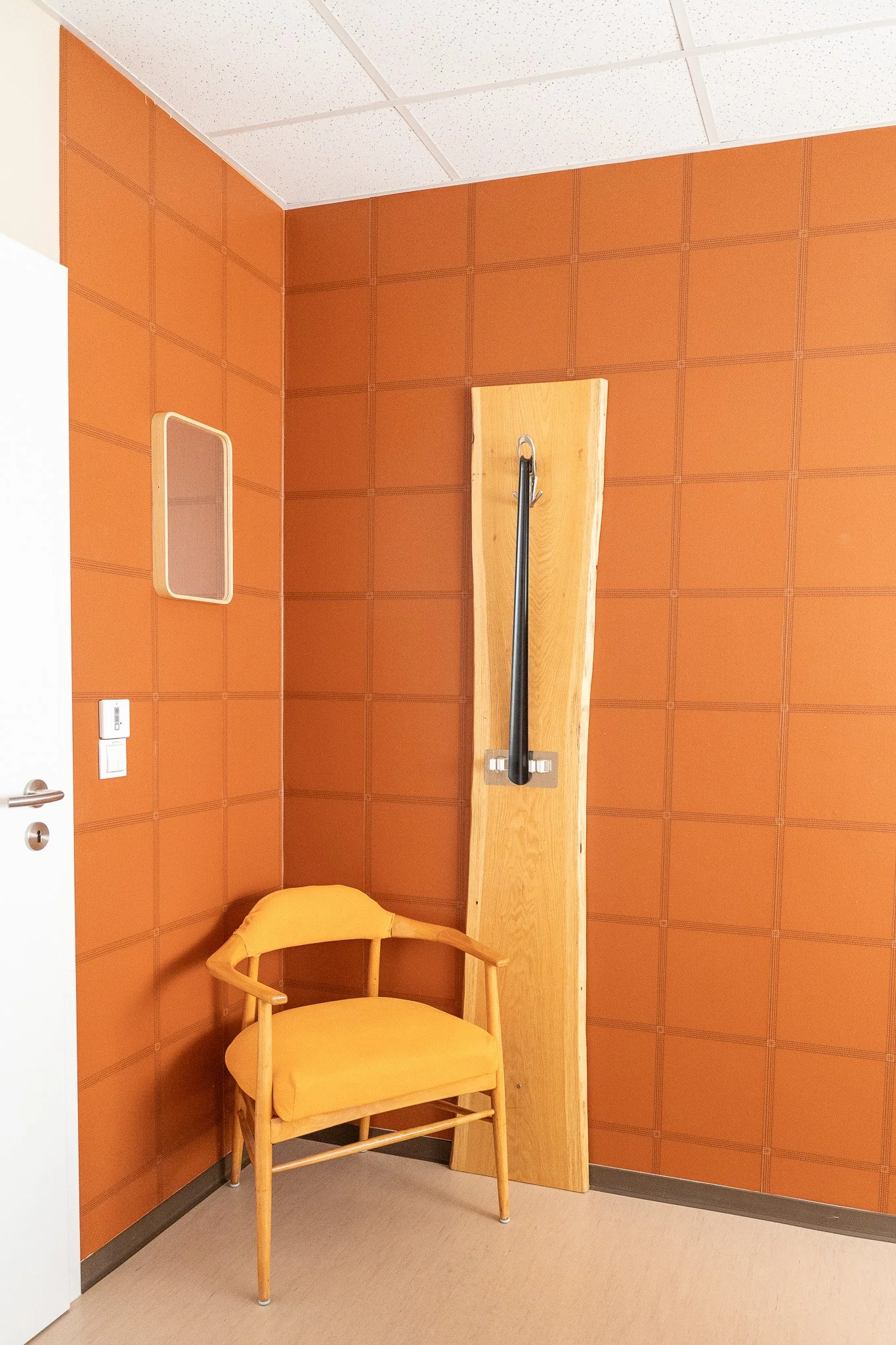 An orange padded wall with a wooden chair and a wall-mounted wooden plank with a metal handrail in an interior room.