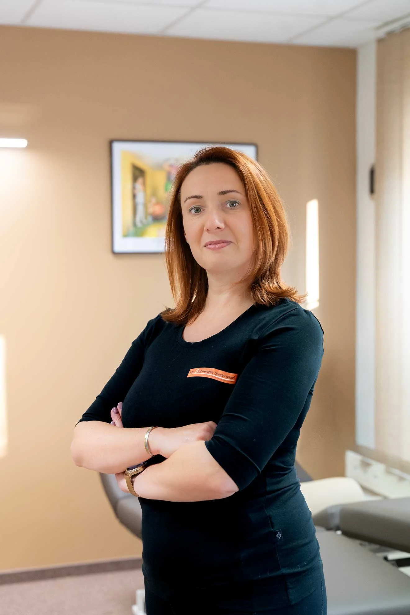 A woman with shoulder-length red hair standing with arms crossed in a room with beige walls and a framed colorful picture in the background.