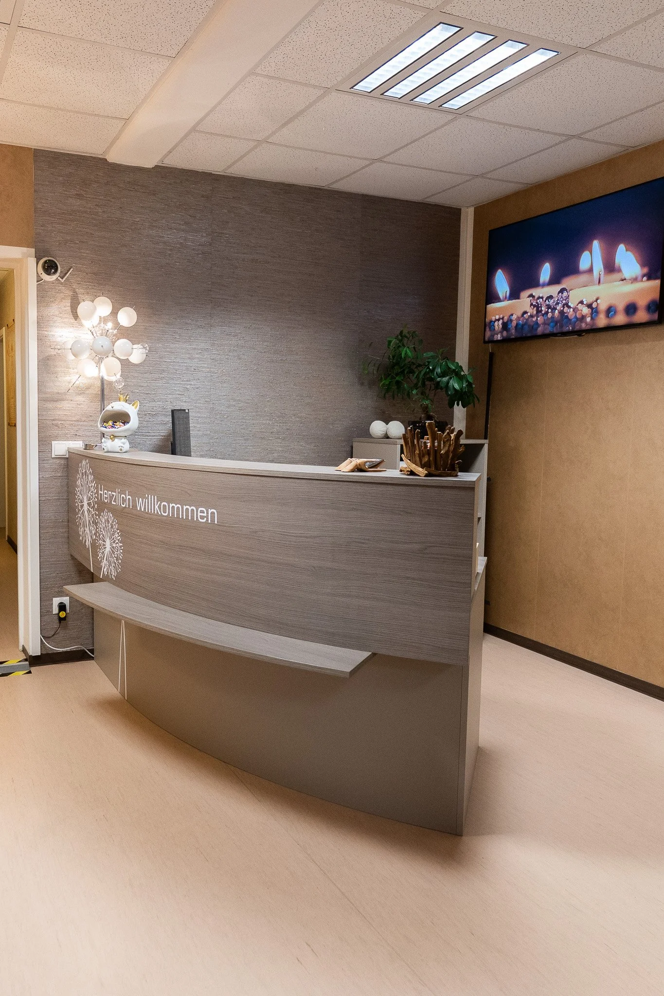 Hospital or clinic reception desk in modern design, with a gray and beige color scheme, a small potted plant, decorative objects, a wall-mounted screen showing candles, and greeting text in German that says 'Herzlich willkommen'.
