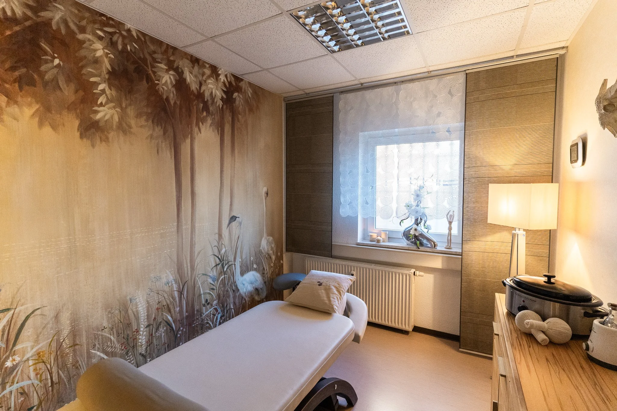 A room with a mural of trees and reeds with swans on a wall, a massage table with a pillow, a window with sheer curtains, a small table with candles, a lamp, a piece of equipment, and a radiator.